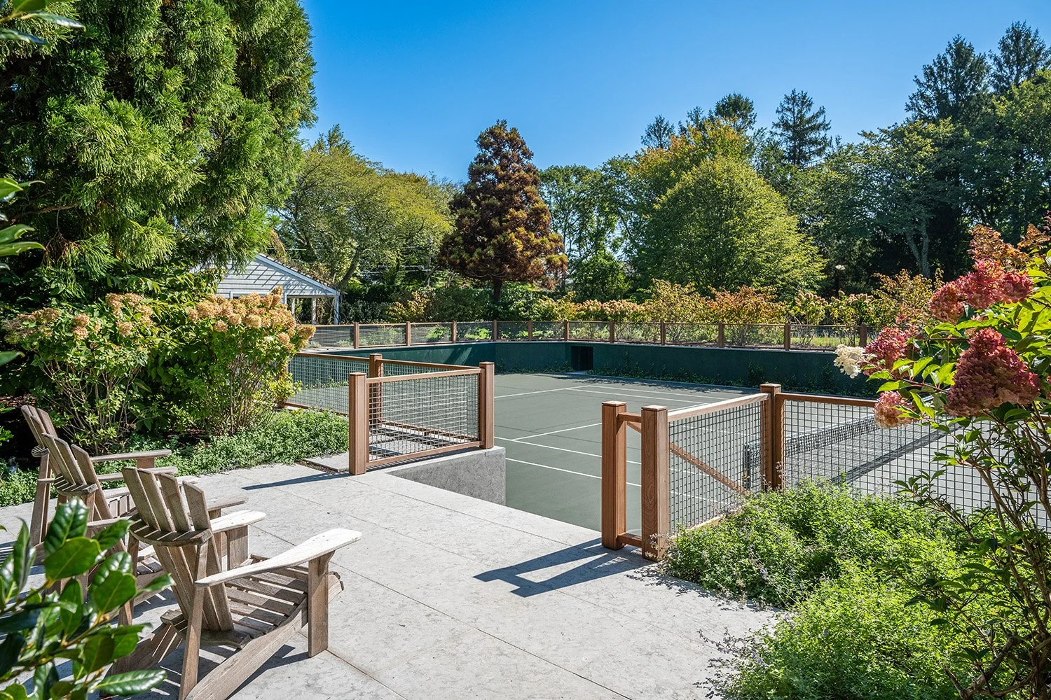 A backyard scene with a tennis court surrounded by lush green trees and blooming flowers, with wooden chairs on a concrete patio in the foreground.