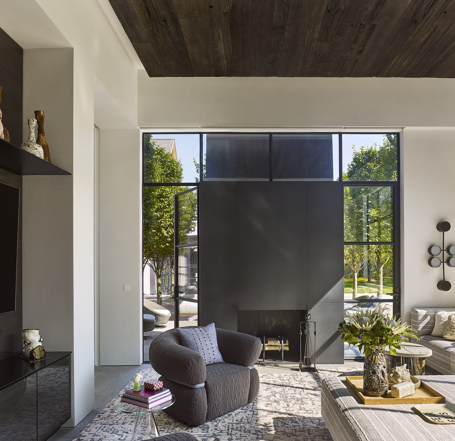 Modern living room with large black fireplace, gray armchair with pillow, patterned rug, glass coffee table with books and a vase of flowers, and large windows showing trees and outdoor seating.