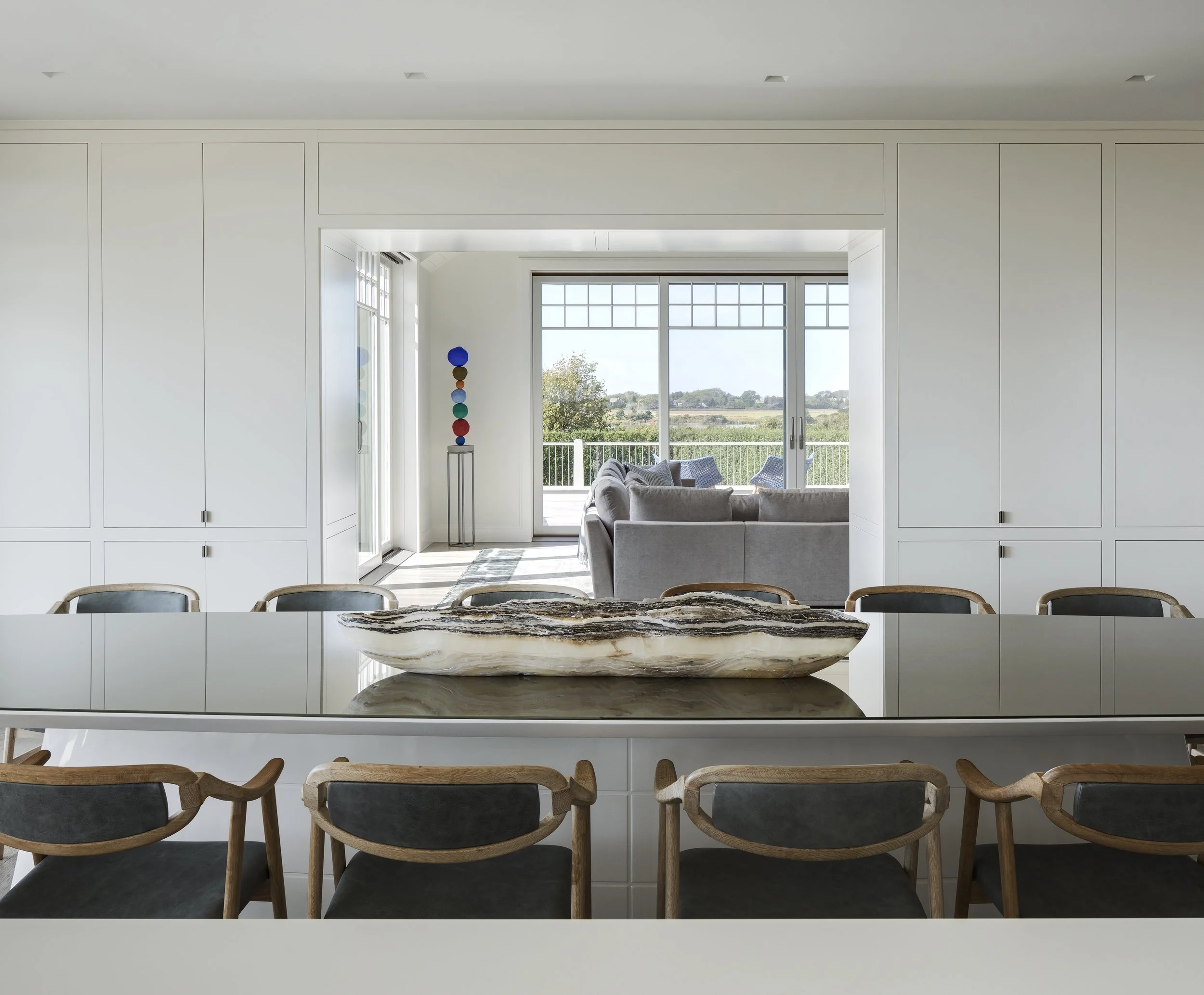 Modern white dining area with a large marble centerpiece, black-cushioned chairs with wooden frames, and a view of a bright living room with a gray sofa and sliding glass doors leading to an outdoor balcony.