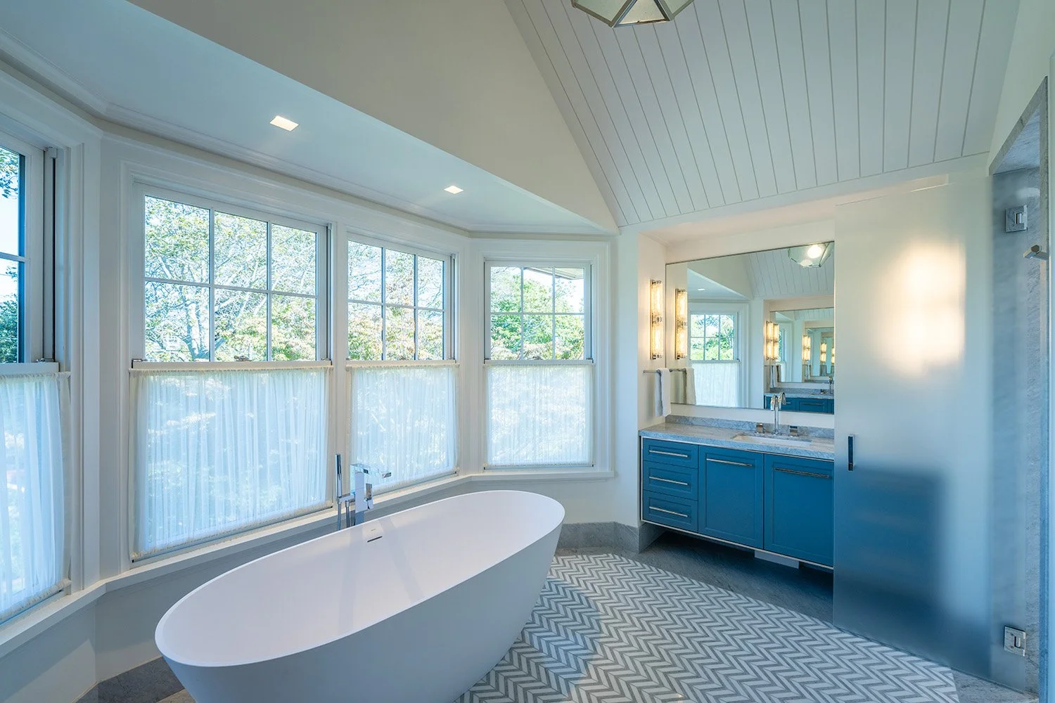 Modern bathroom with white freestanding bathtub, large windows with sheer curtains, blue vanity with granite countertop, and a mirror reflecting the room's light and greenery outside.