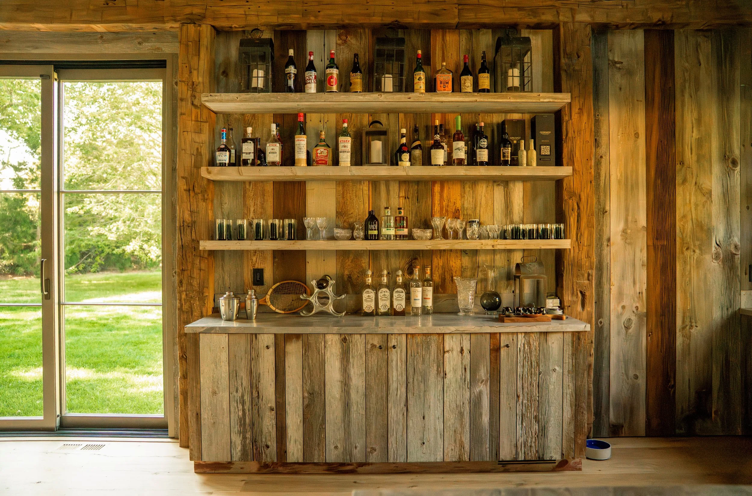 A rustic wooden home bar with shelves holding bottles of alcohol and glasses, next to a glass door opening to a green outdoor space.