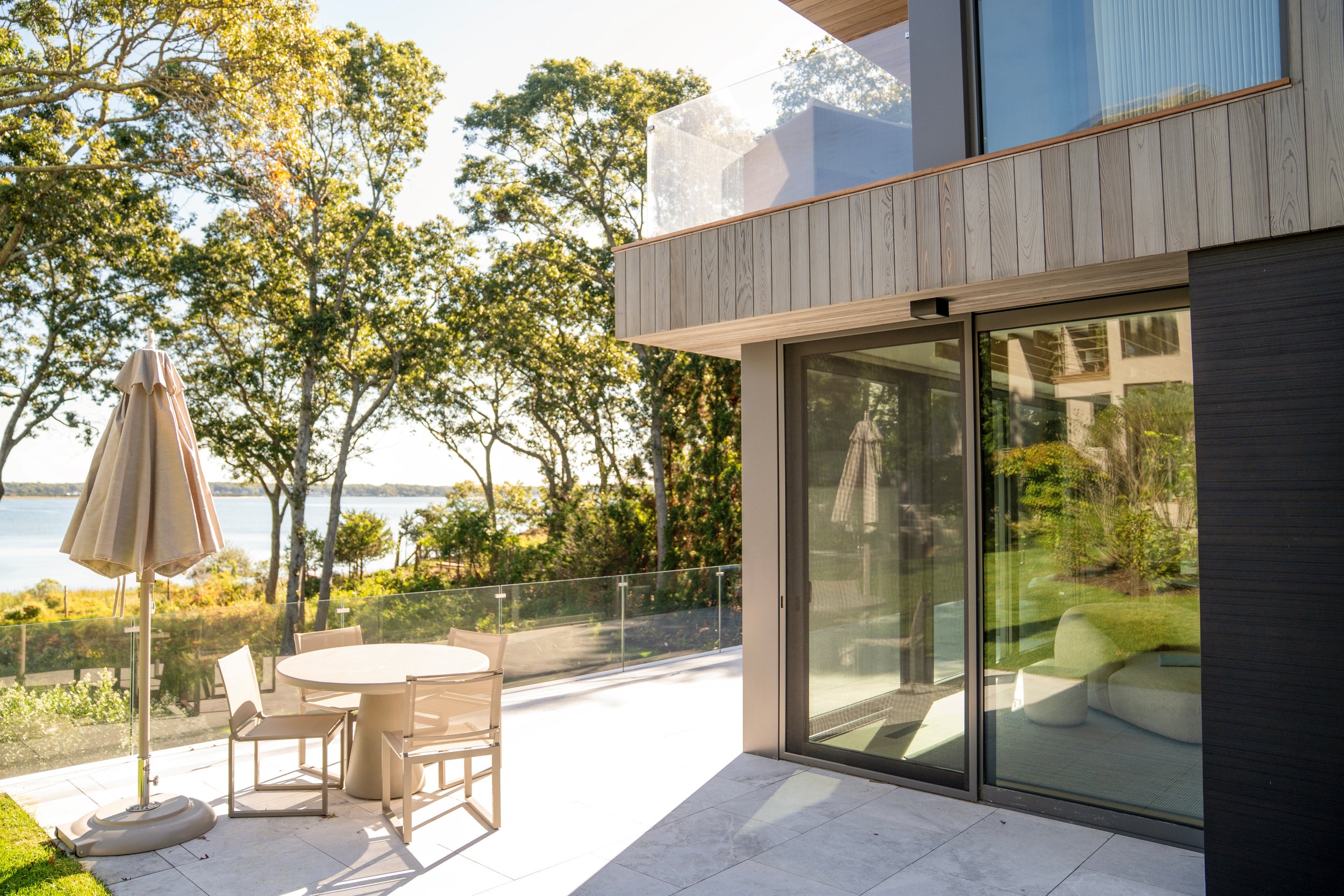 Modern house with large glass sliding doors, outdoor patio with dining table, chairs, and umbrella, overlooking a lake and wooded area.