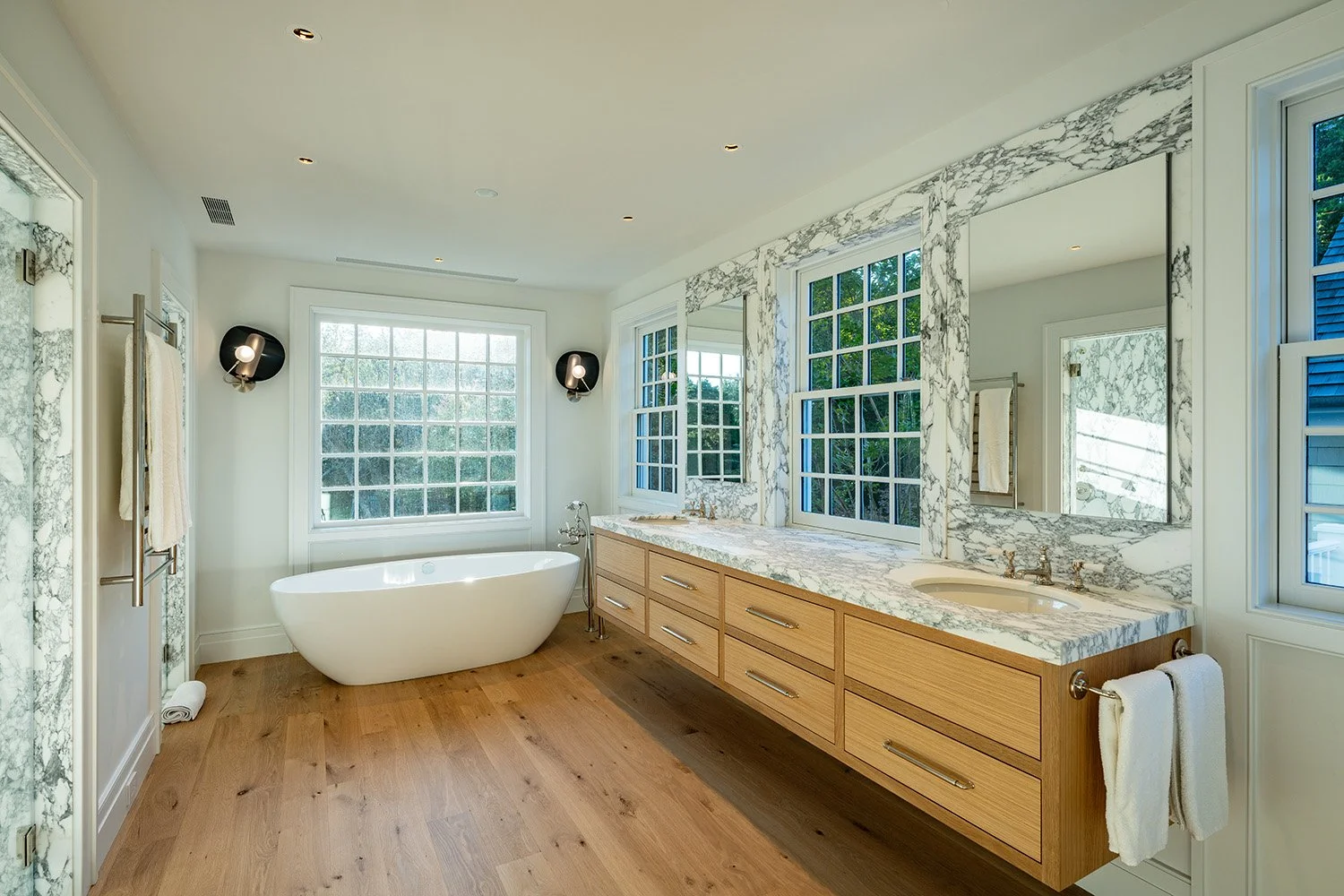 Luxury bathroom featuring a freestanding bathtub, marble countertop with a sink, large windows with grid panes, wood flooring, and modern wall-mounted lights.