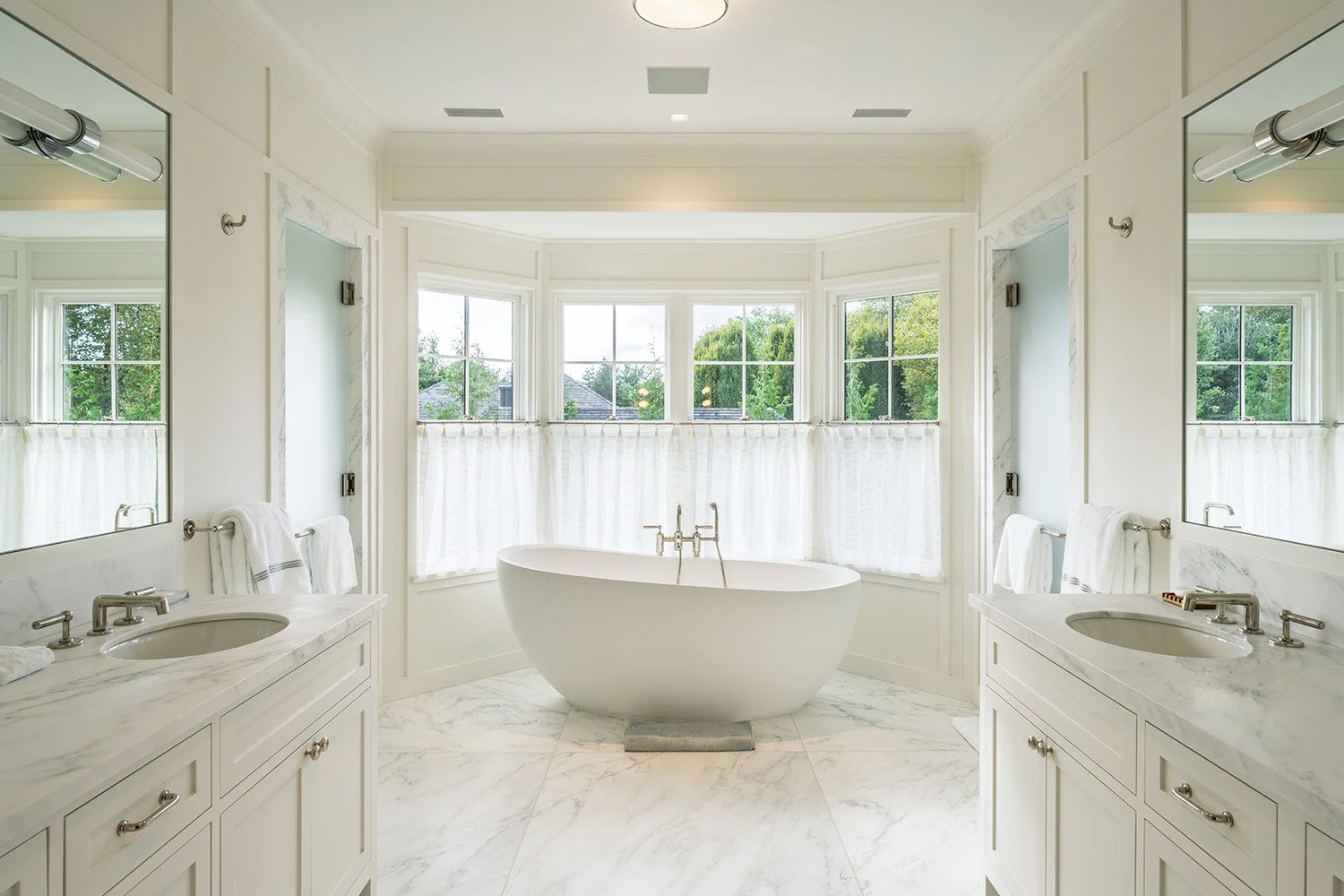 Bright and spacious bathroom with a freestanding bathtub centered in front of a large bay window with white curtains. The bathroom features two white marble vanities with sinks, mirrors, and towels, with greenery visible through the windows.