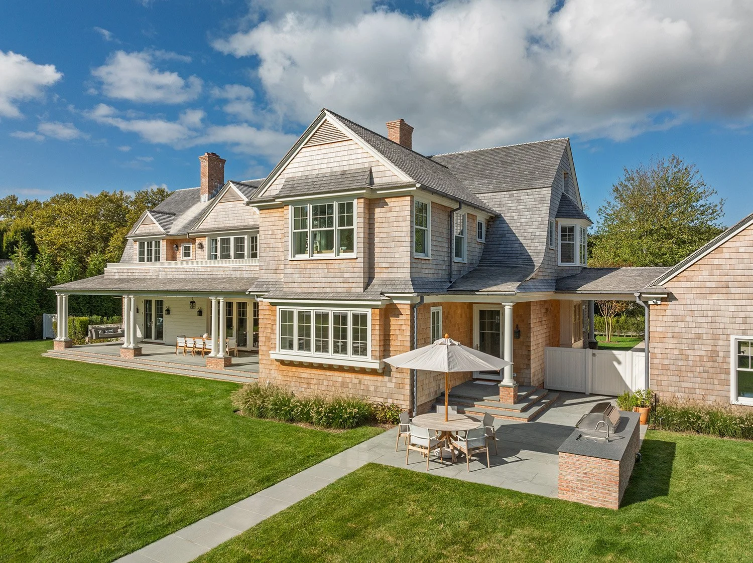 Large, multi-story house with a brick and shingle exterior, multiple windows, and a spacious backyard patio with outdoor furniture and an umbrella. Construction in Hamptons New York