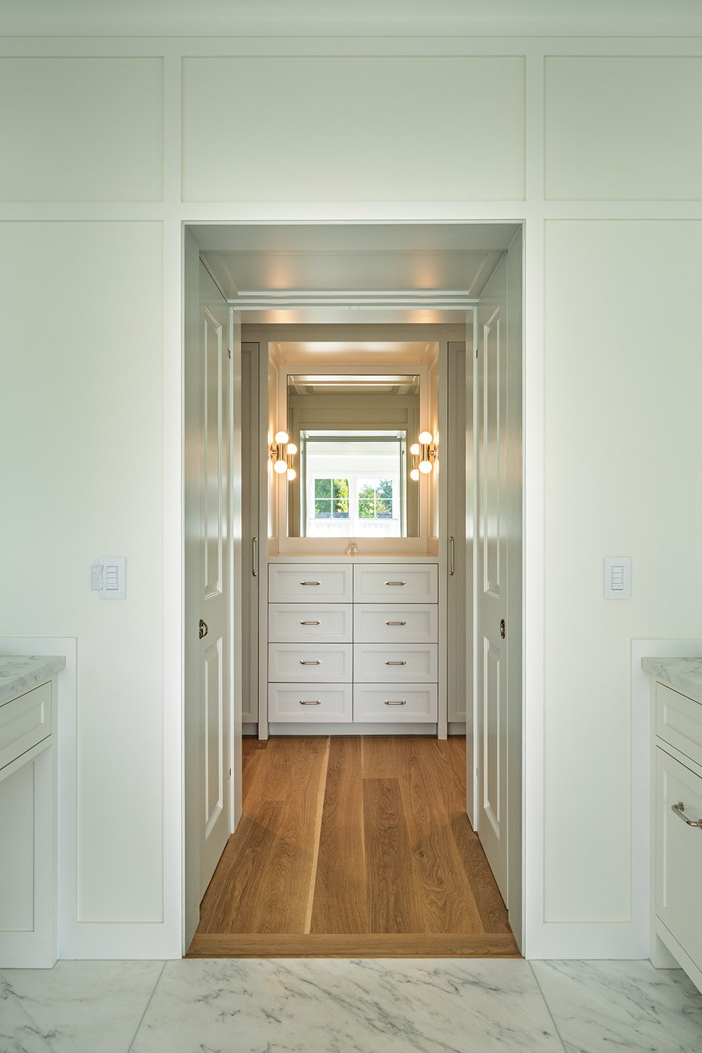 View through a doorway into a bright dressing room with white cabinetry, drawers, a large mirror, and modern light fixtures, with windows showing greenery outside.