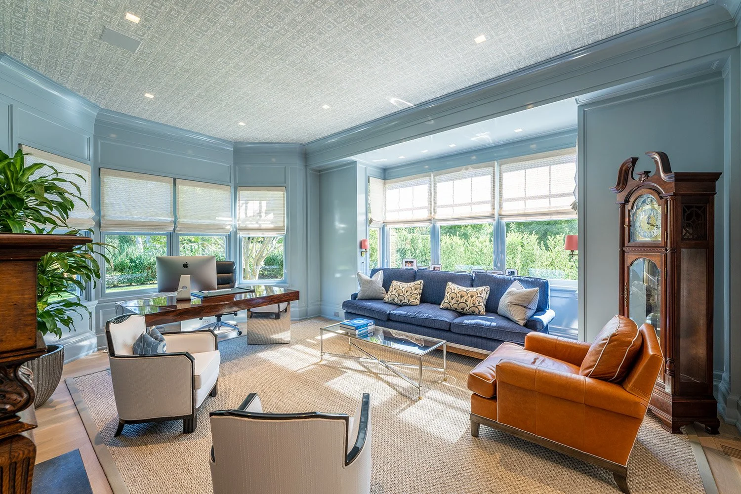 Bright living room with blue walls, large windows, blue sofa with decorative pillows, orange armchair, glass coffee table, vintage wooden grandfather clock, white armchairs, and a desk with a computer, decorated with houseplants and framed photos.