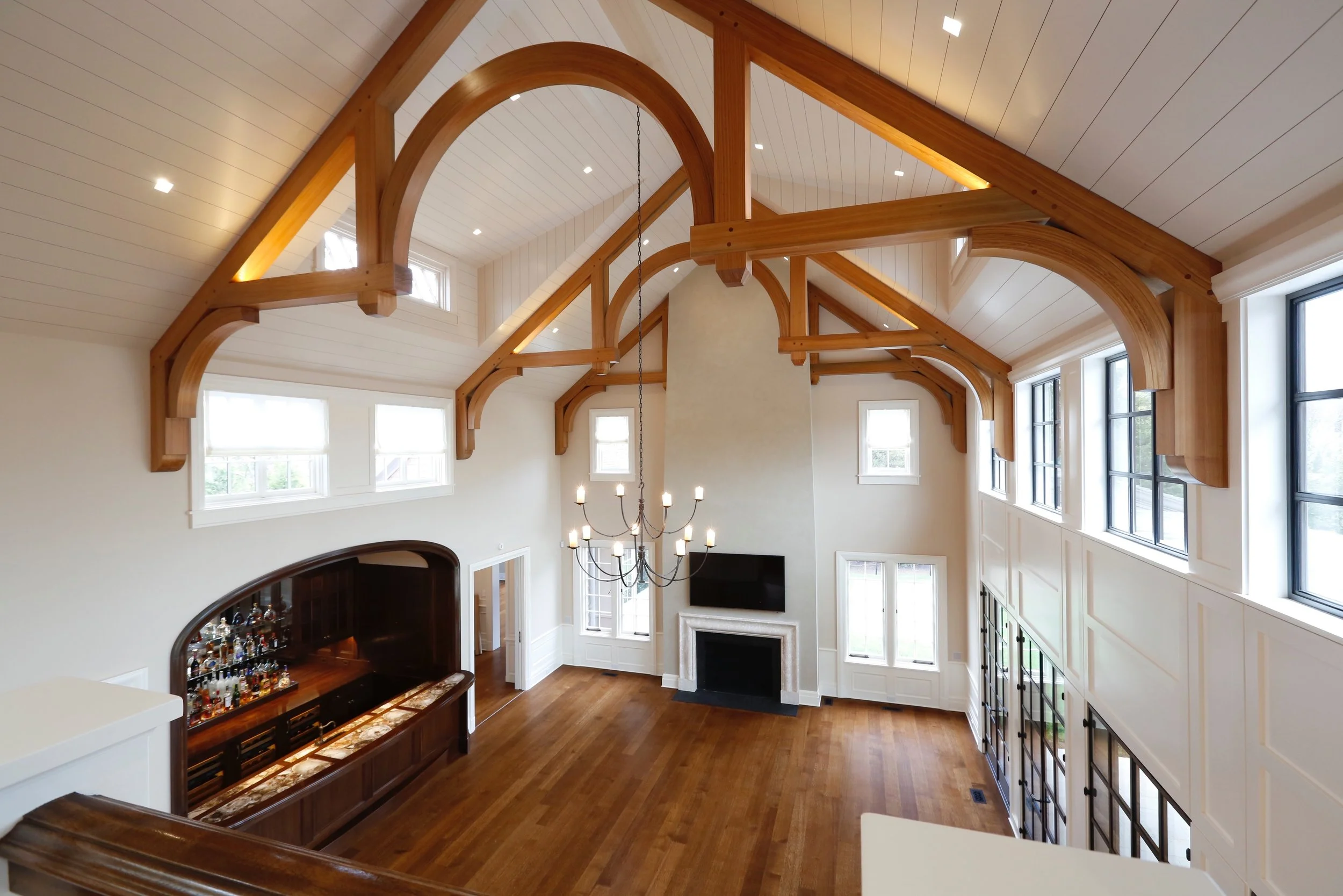 Interior of a spacious living room with a vaulted ceiling, exposed wooden beams, large windows, a chandelier, a fireplace, and a built-in bar area.