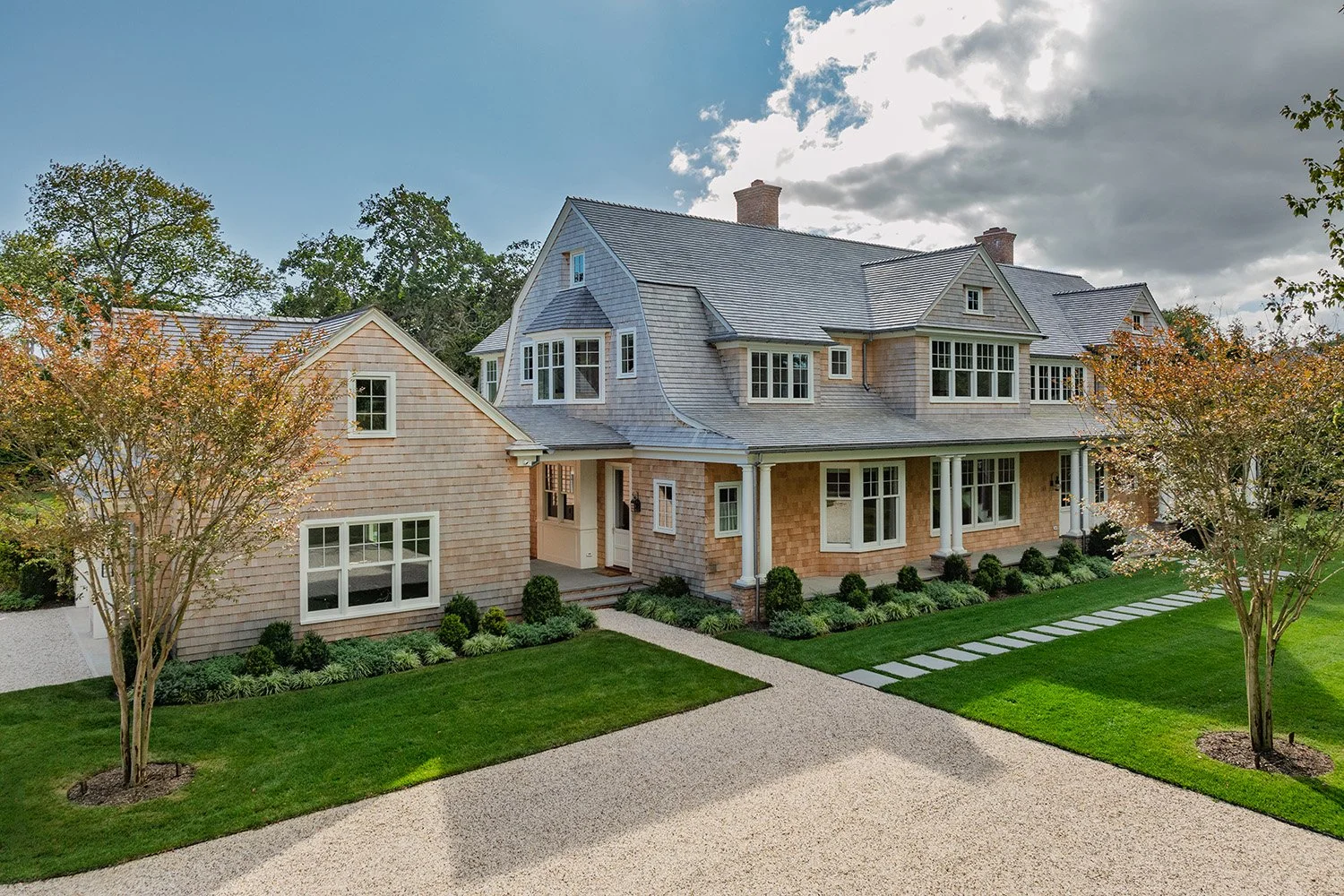 Large two-story house with shingle siding, multiple windows, a covered porch with white columns, a gravel driveway, and a landscaped front yard with trees, bushes, and a walking path. Construction in Hamptons New York