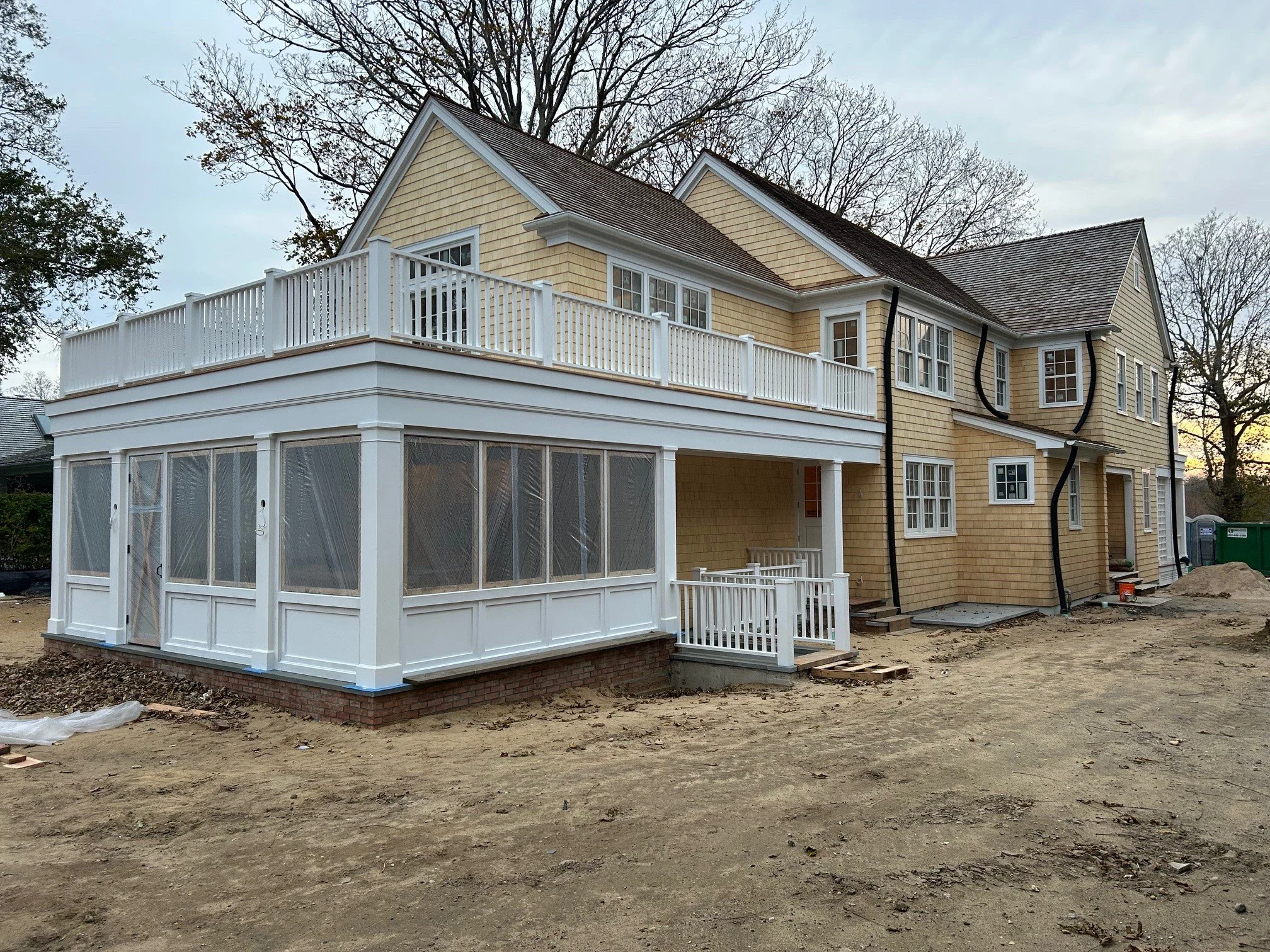 A two-story house under construction with a yellow exterior, white trim, and a large deck with white railings on the second floor. The house has multiple windows and a screened-in porch on the ground floor. The yard is unfinished with dirt and constr
