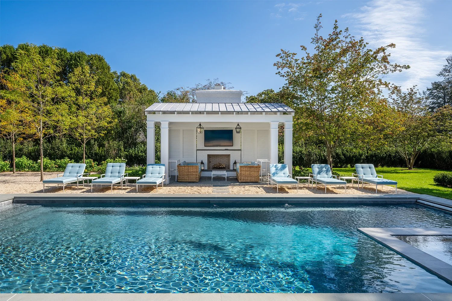 Luxury backyard with swimming pool, white lounge chairs, outdoor fireplace, flat-screen TV, and trees in the background under a blue sky.