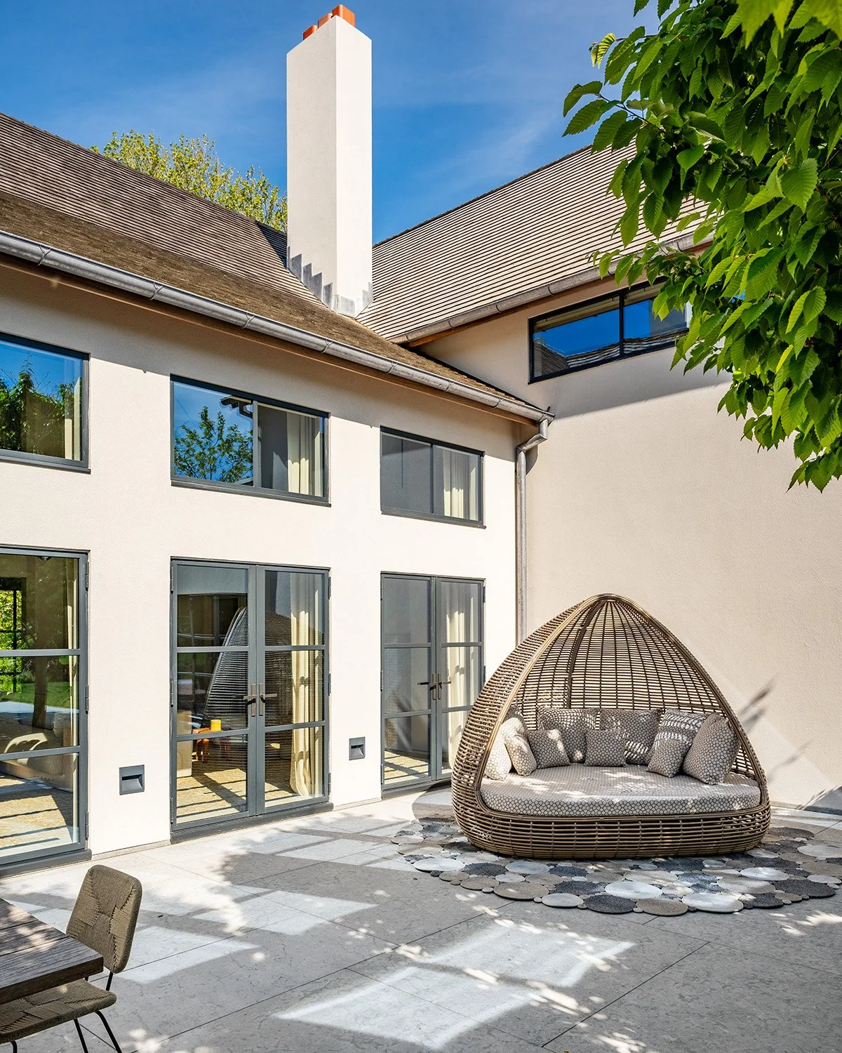Modern house with large windows, a covered outdoor seating area with cushions in a woven dome-shaped structure, surrounded by greenery and a clear blue sky.