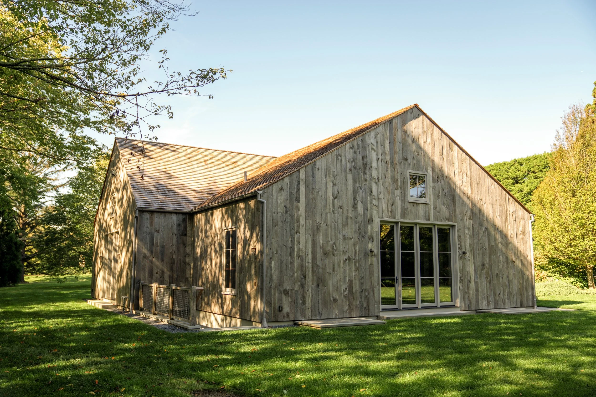 A modern house with a wooden exterior, large glass doors, and a yard with grass and trees, under a clear sky.