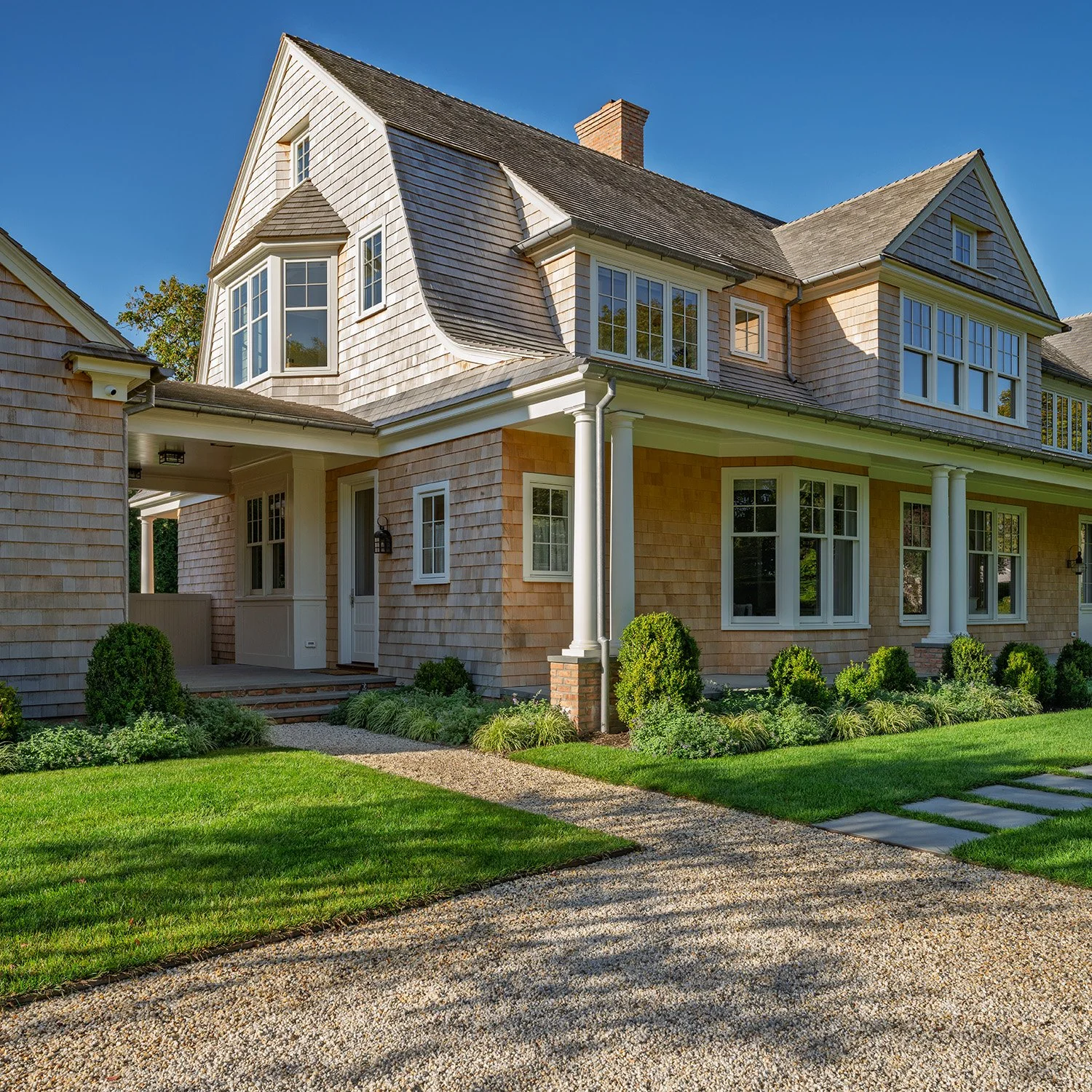 A large, two-story house with beige wooden siding, multiple windows, and a front porch with columns, surrounded by a green lawn and garden. Construction in Hamptons New York