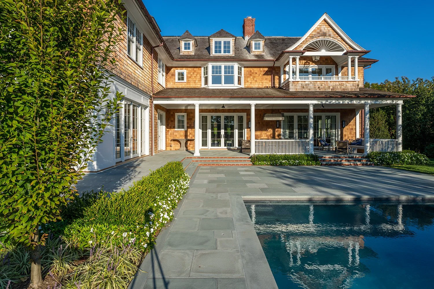 Large backyard with a swimming pool, landscaped garden with bushes and trees, outdoor seating area, and a multi-story house with a porch, large windows, and a gabled roof.