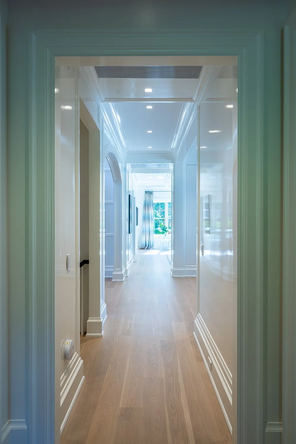 Bright hallway with white walls, crown molding, and wooden flooring, leading to a sunlit room with large window and curtains.