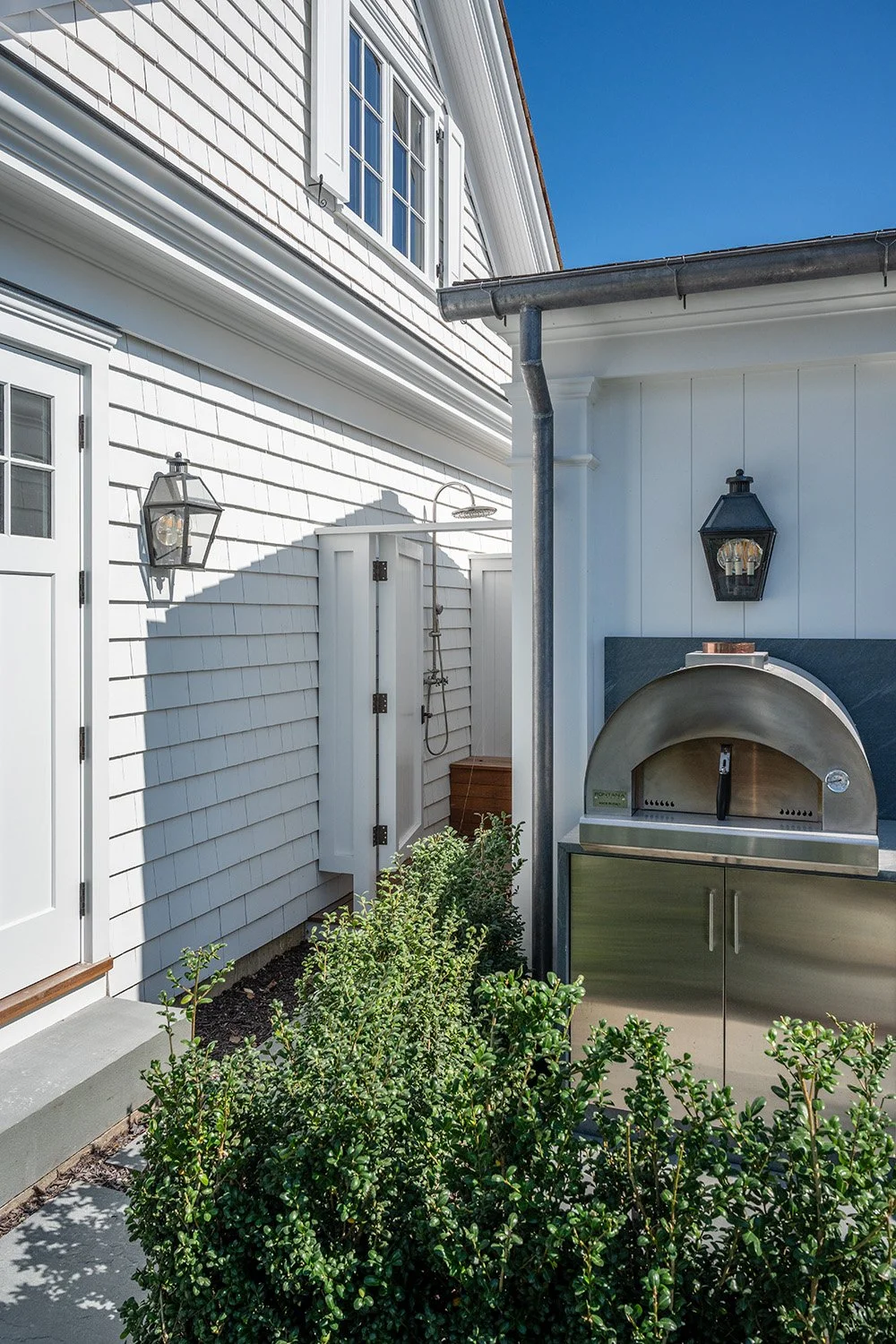 Outdoor scene of a white house with a door, outdoor shower, and a stainless steel pizza oven, with greenery and a clear blue sky.