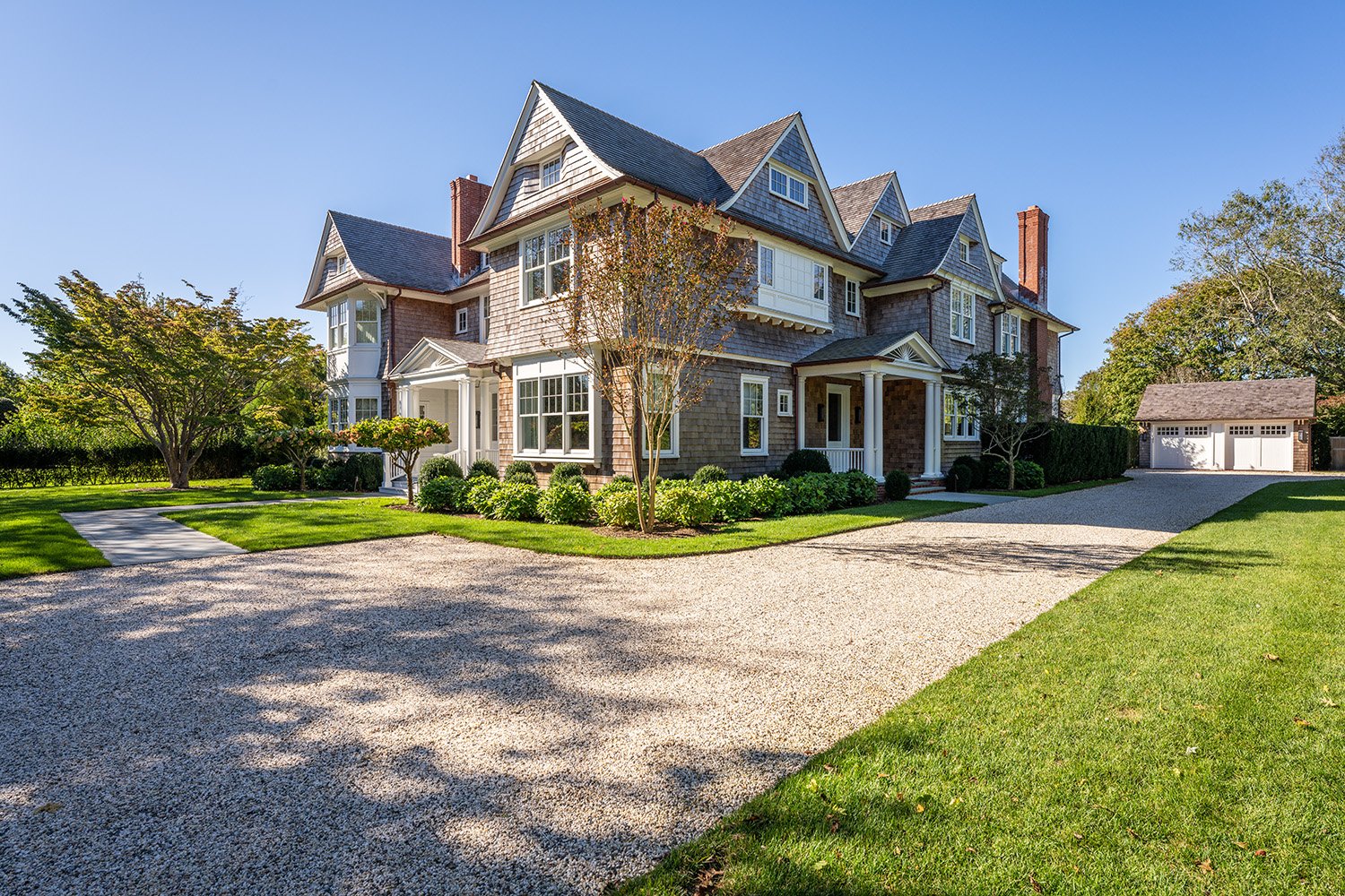 Large Victorian-style house with multiple gables and a front porch, surrounded by a well-maintained lawn and trees under a clear blue sky.