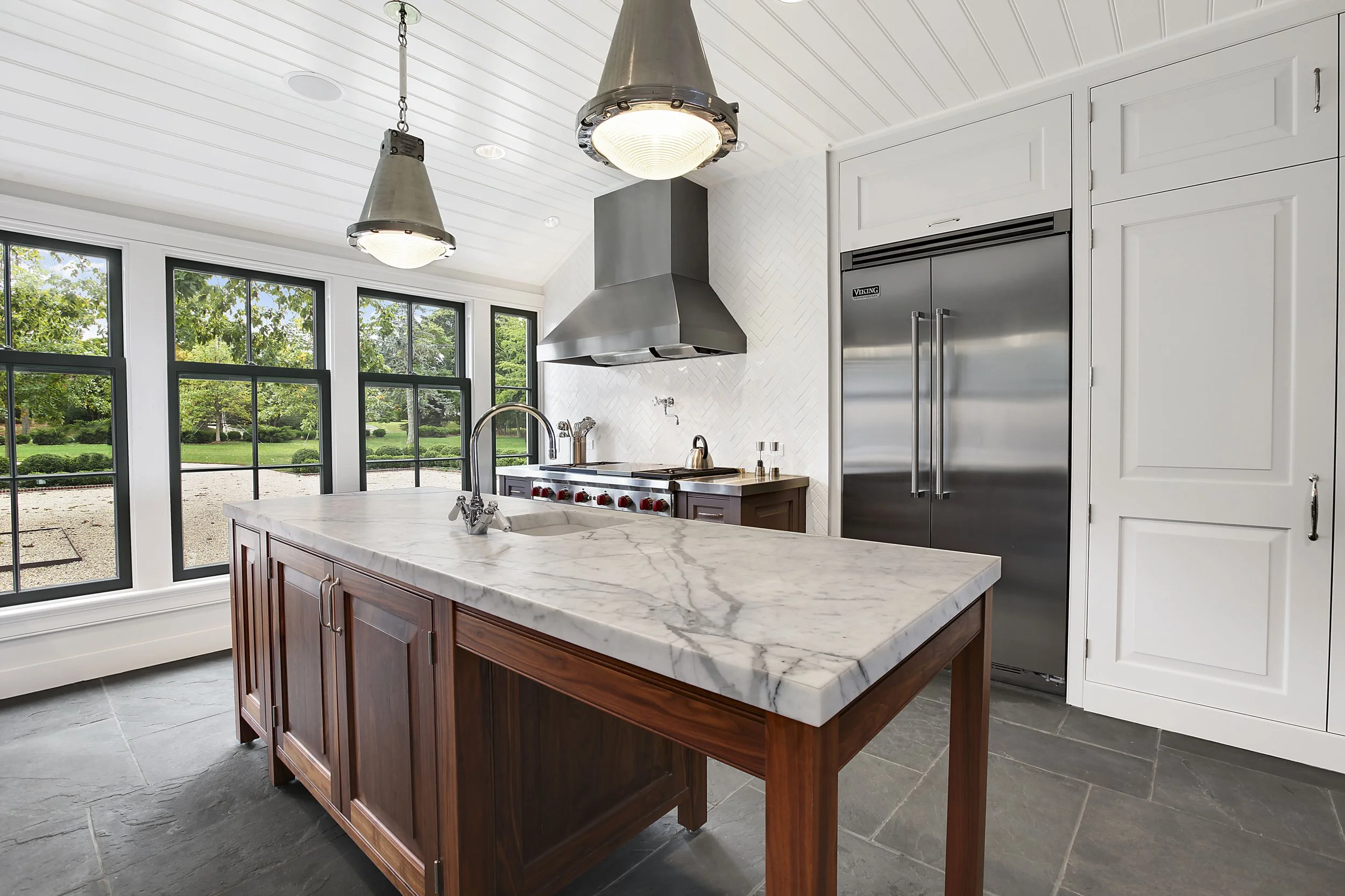 Modern kitchen with marble island, stainless steel refrigerator, large windows, and pendant ceiling lights.