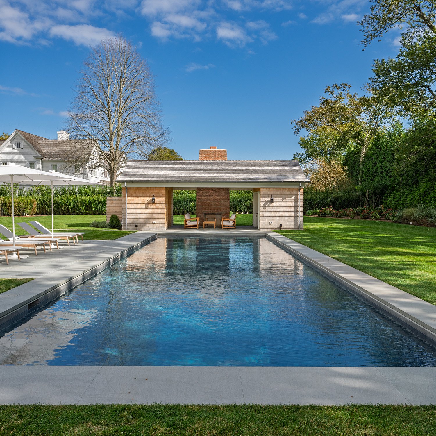 A backyard with a swimming pool, surrounded by a grassy lawn, patio chairs, and a small pavilion with outdoor seating and a brick chimney.