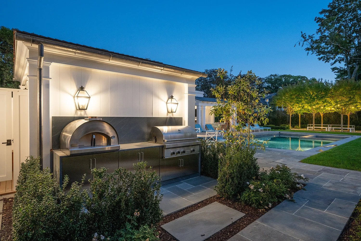 Backyard with outdoor kitchen featuring a pizza oven and grill, a swimming pool, lounge chairs, trees, and landscaped garden at dusk.