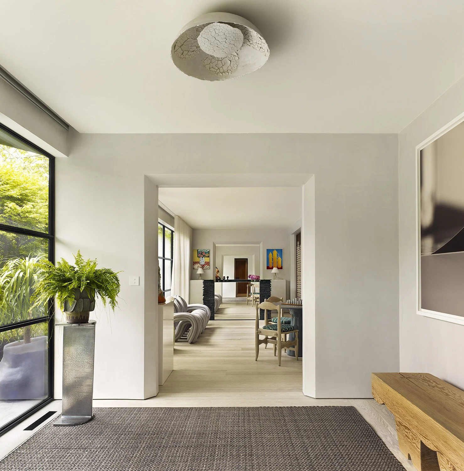 View through an entryway into a living room with white walls, wooden flooring, potted plant on a stand, and artwork on the walls.