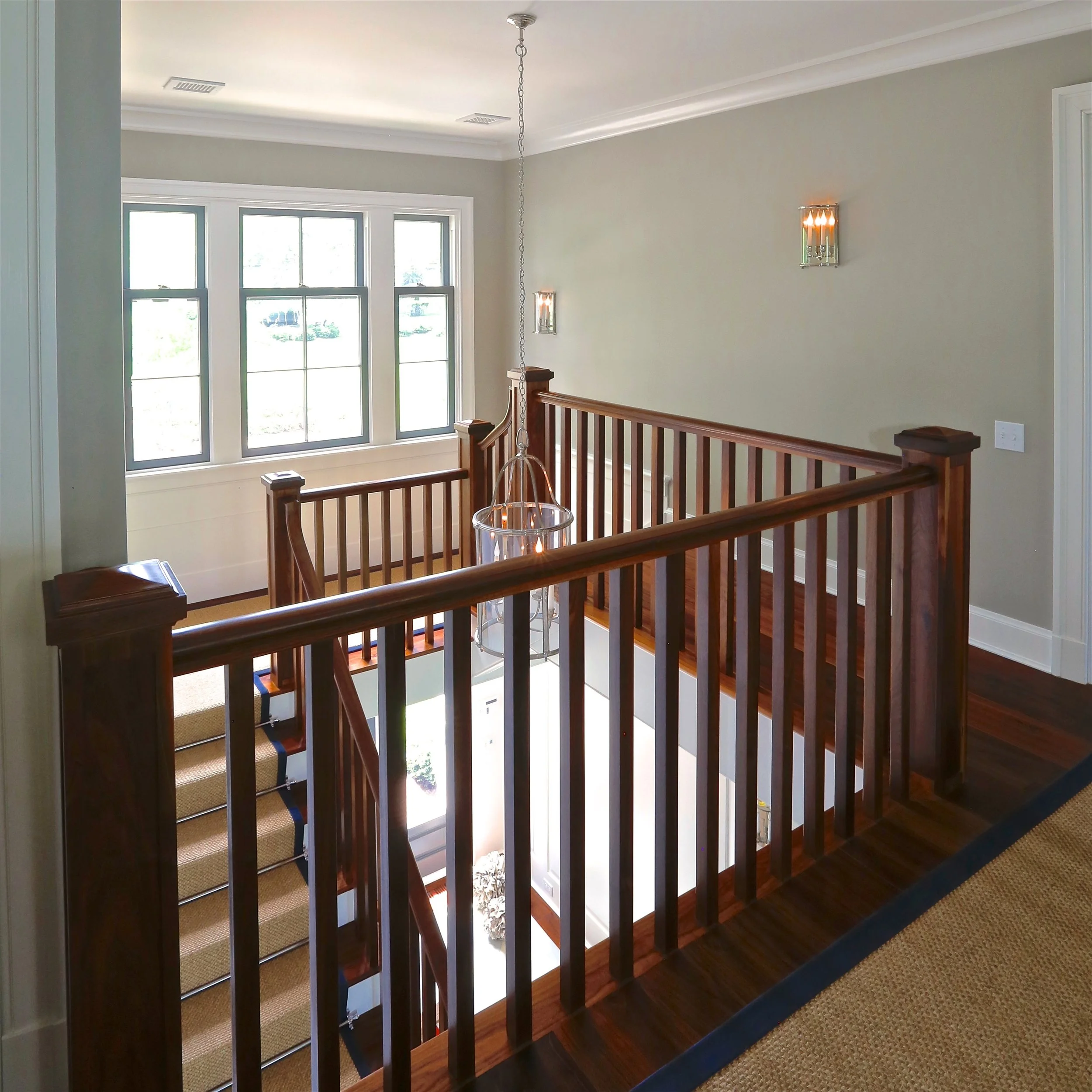 Interior view of a house with a staircase, wooden banister, large window, chandelier, and wall sconces.