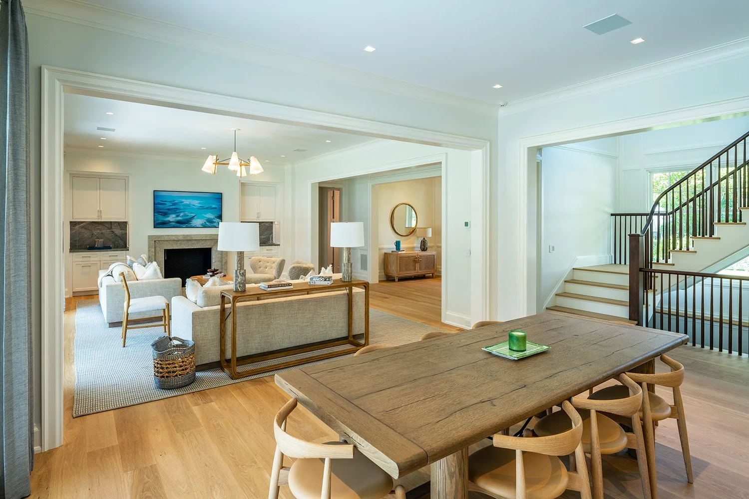 Open-concept living and dining area with hardwood floors, white walls, and large windows letting in natural light; living room has a sofa and armchair with a fireplace and a wall-mounted TV; dining area features a rustic wooden table with chairs, and