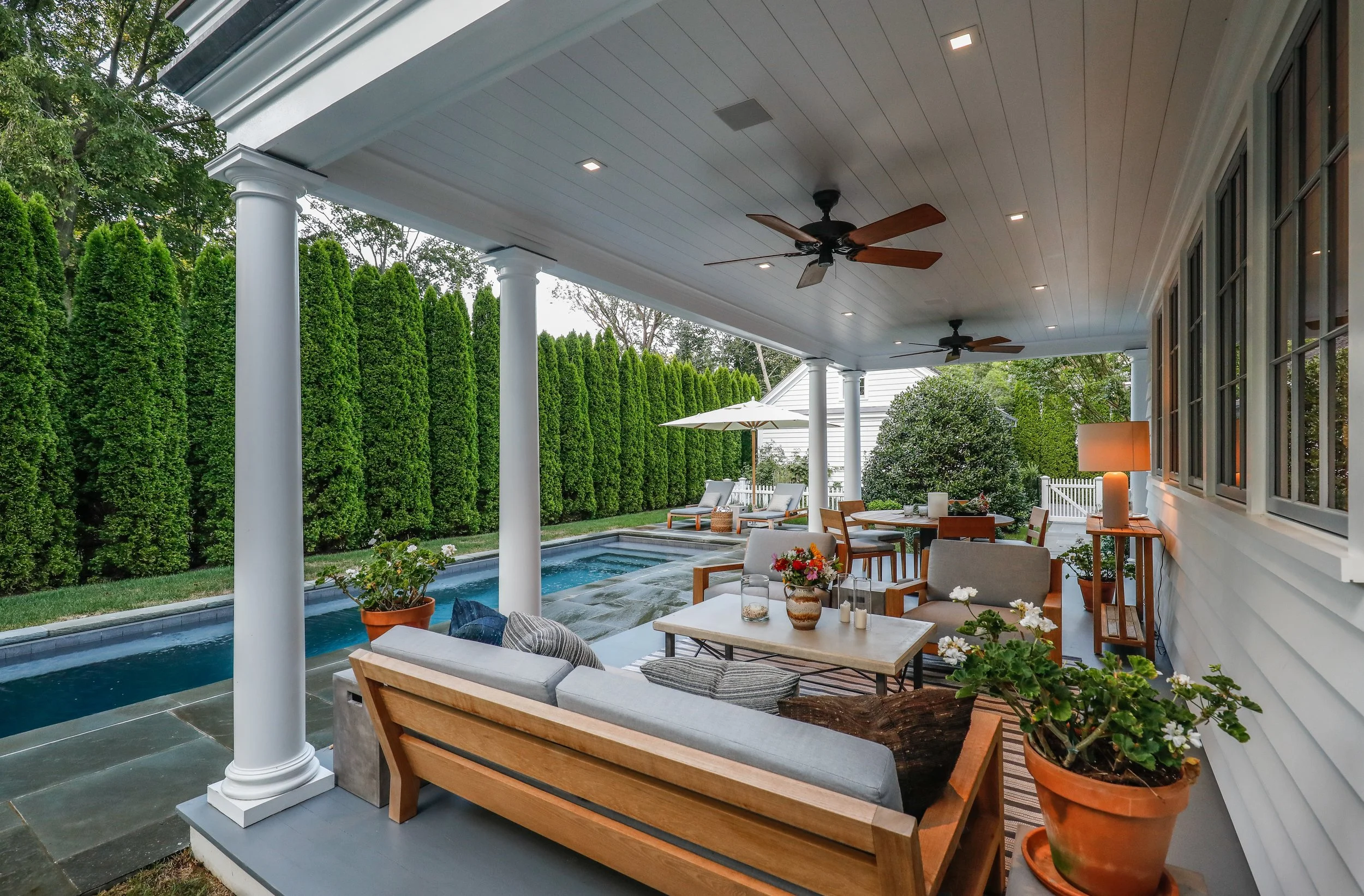 Covered backyard patio with outdoor furniture, potted plants, and a small swimming pool, surrounded by tall green hedges. Construction in Hamptons New York