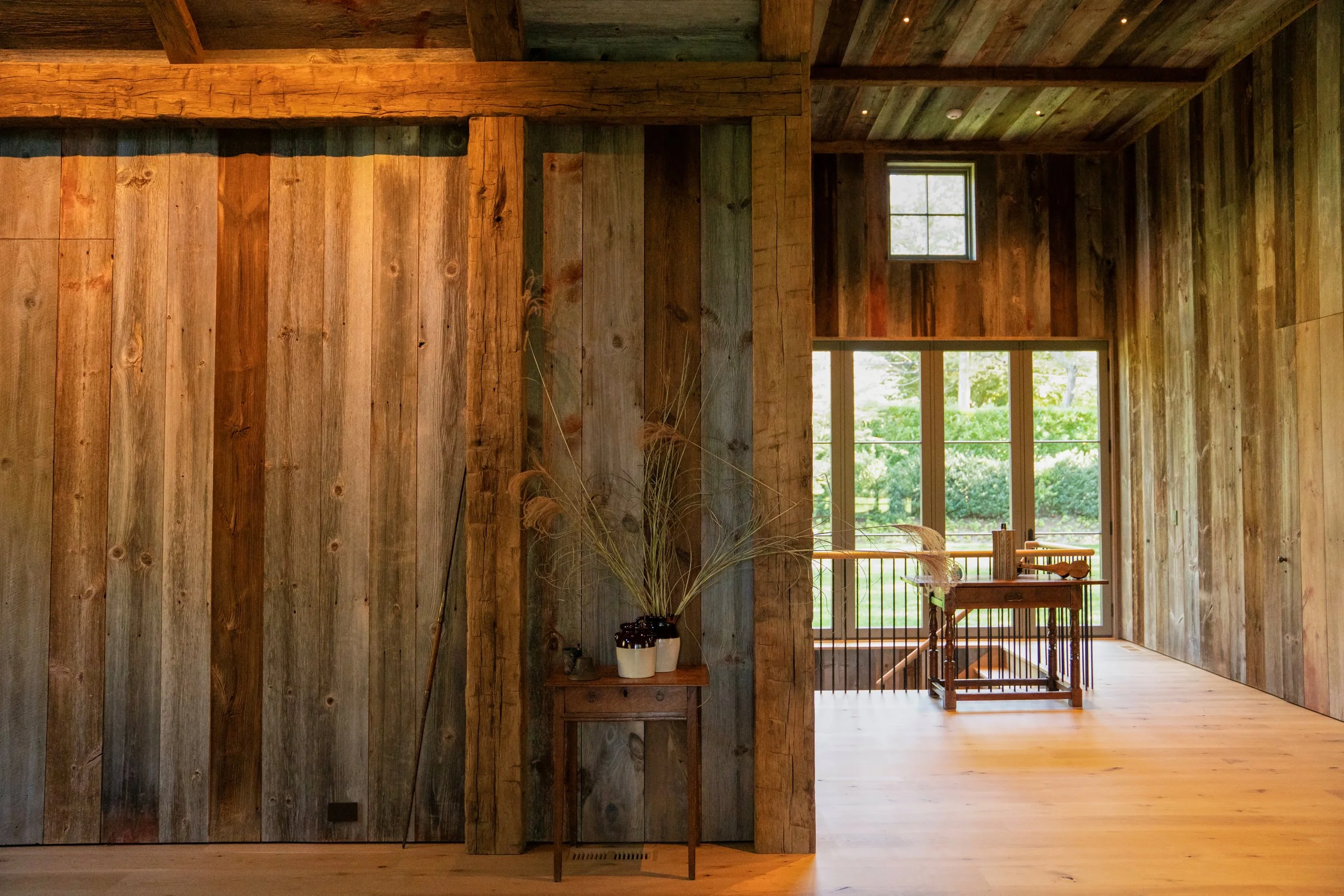 Interior of a rustic wooden house featuring vertical reclaimed wood paneling, a small table with potted plants, and a large window showing greenery outside. Custom residential construction building in the Hamptons
