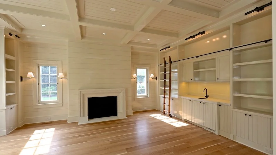 Empty room with white walls, hardwood floors, two windows, built-in shelves, a fireplace, and a ceiling with exposed beams. There is a collapsible ladder and a long curtain rod along the wall. Amagansett construction