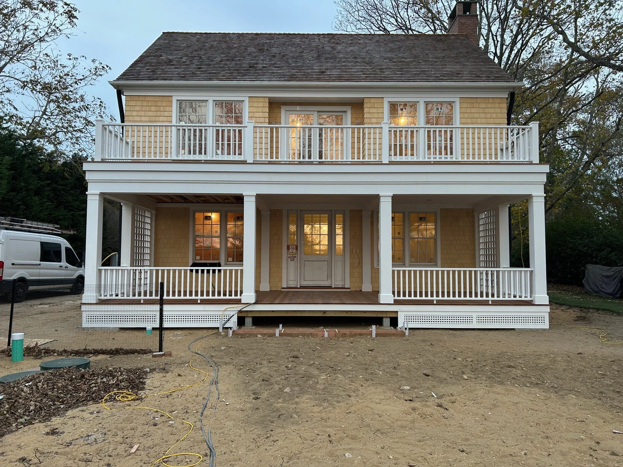 A newly constructed two-story house with yellow siding, white trim, and a large front porch with railings, set on a dirt yard with construction wires and equipment visible. Sag Harbor construction