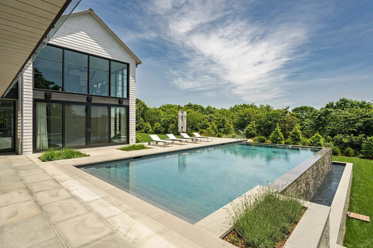 A modern house with large glass windows and patio doors, a rectangular swimming pool with a stone waterfall feature, surrounded by lounge chairs and umbrellas, overlooked by lush green trees and a partly cloudy sky.
