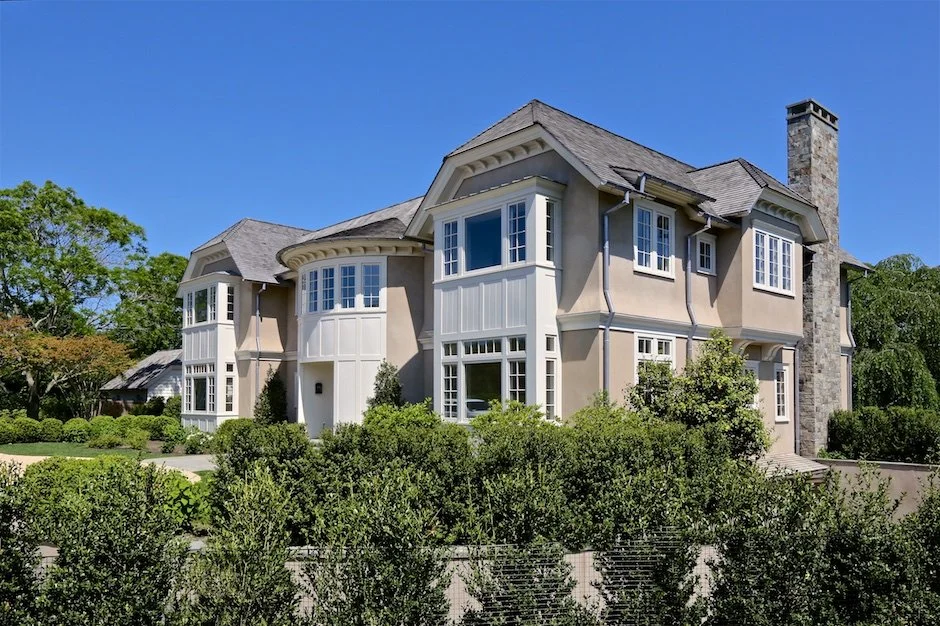 A large beige two-story house with multiple bay windows, a stone chimney, and surrounded by greenery under a clear blue sky. Montauk builders.
