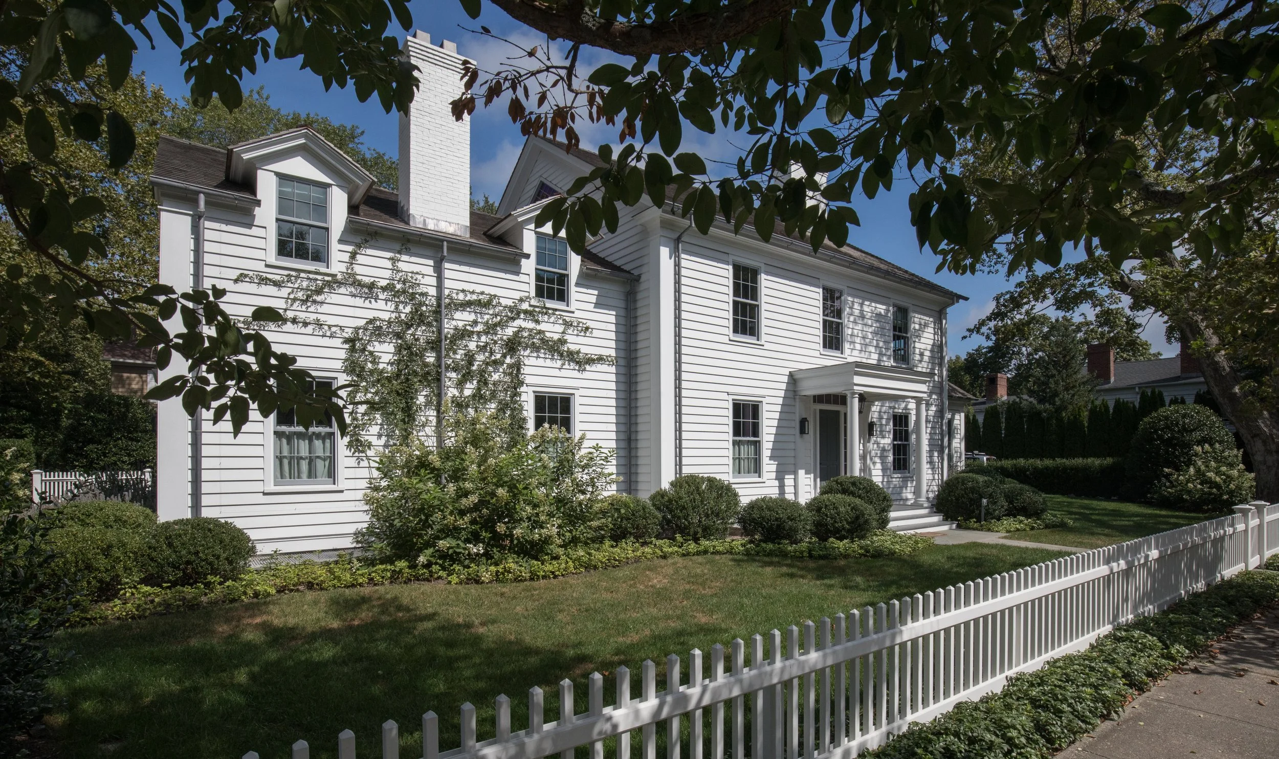 White two-story house with black shutters, surrounded by a garden with bushes, trees, and a white picket fence. Sag Harbor construction company