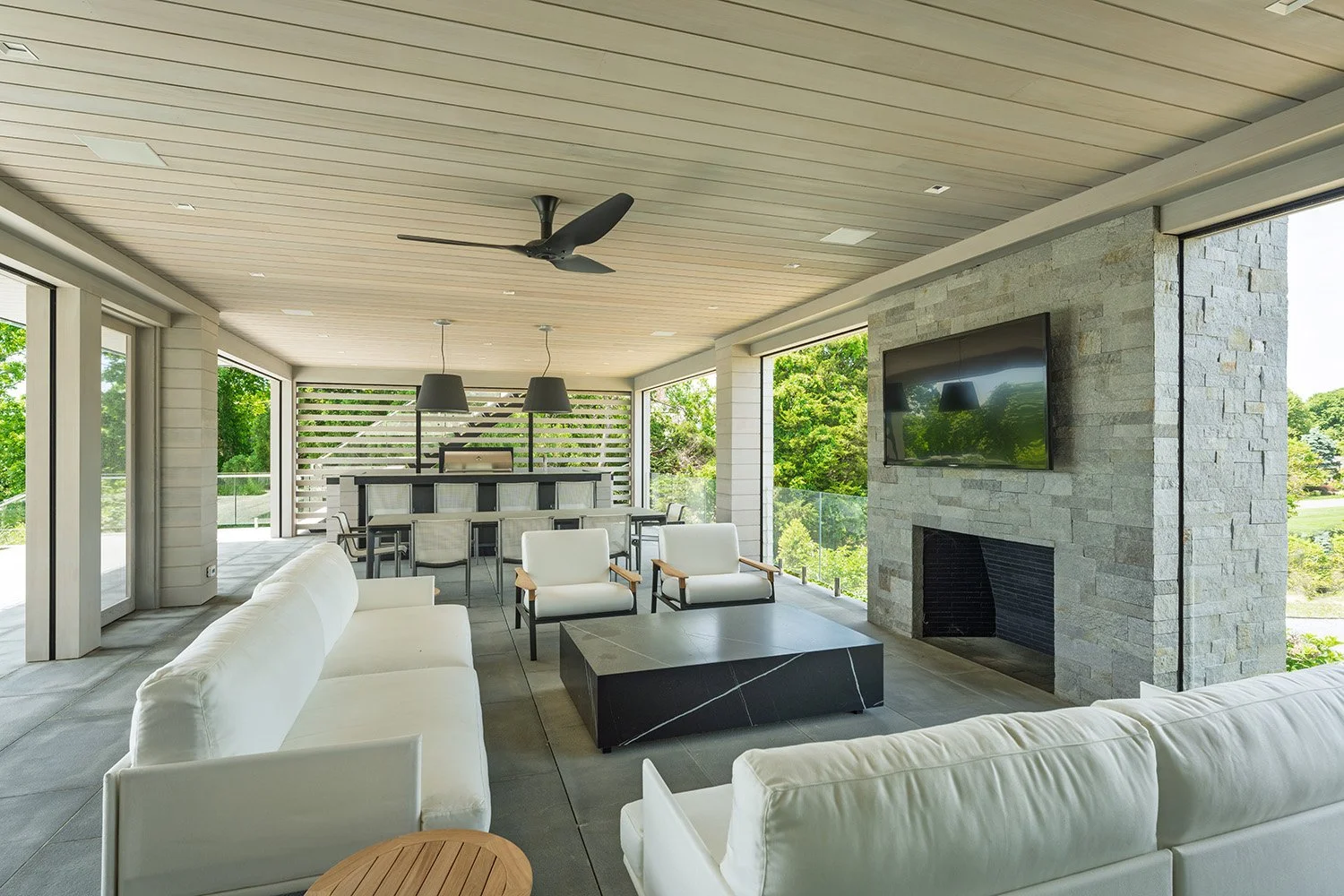 Modern outdoor living space with white sofas, armchairs, a black marble coffee table, a stone fireplace with a mounted TV, a dining table with chairs, large windows, and a ceiling fan, surrounded by lush greenery.