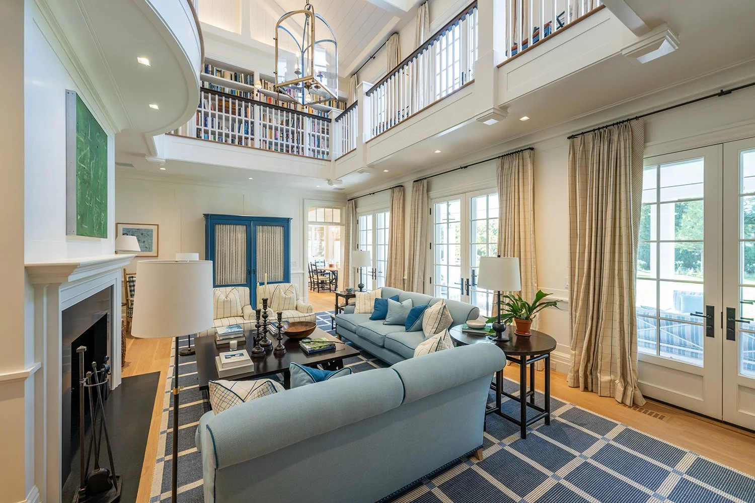 Bright living room with blue and cream furniture, large windows with beige curtains, a fireplace, a black coffee table, and a second-floor balcony filled with bookshelves