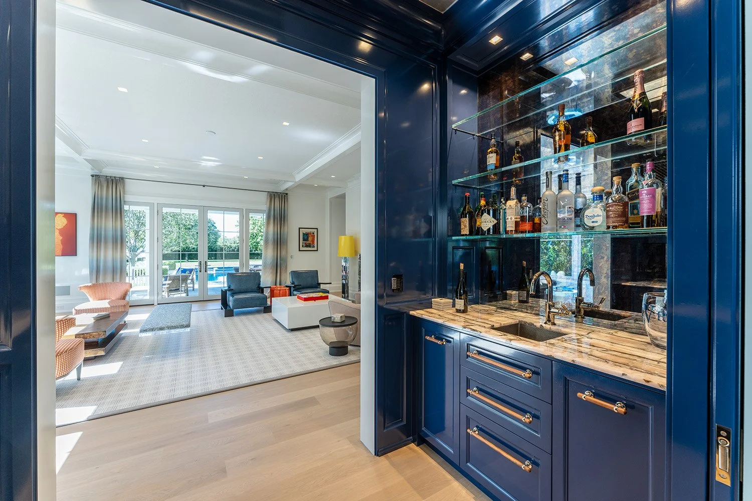 A view from a home bar area showing shelves of liquor bottles behind a blue cabinet with a marble countertop, opening into a bright living room with large windows, sliding doors, and outdoor pool area.