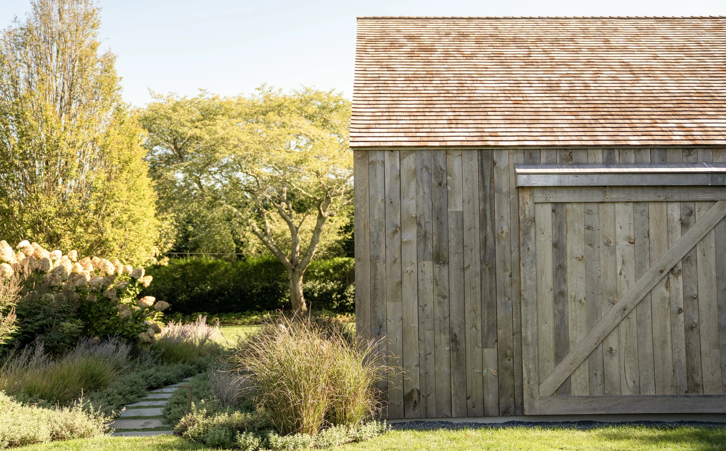 A wooden shed with a red shingled roof next to a landscaped garden with various plants and trees.