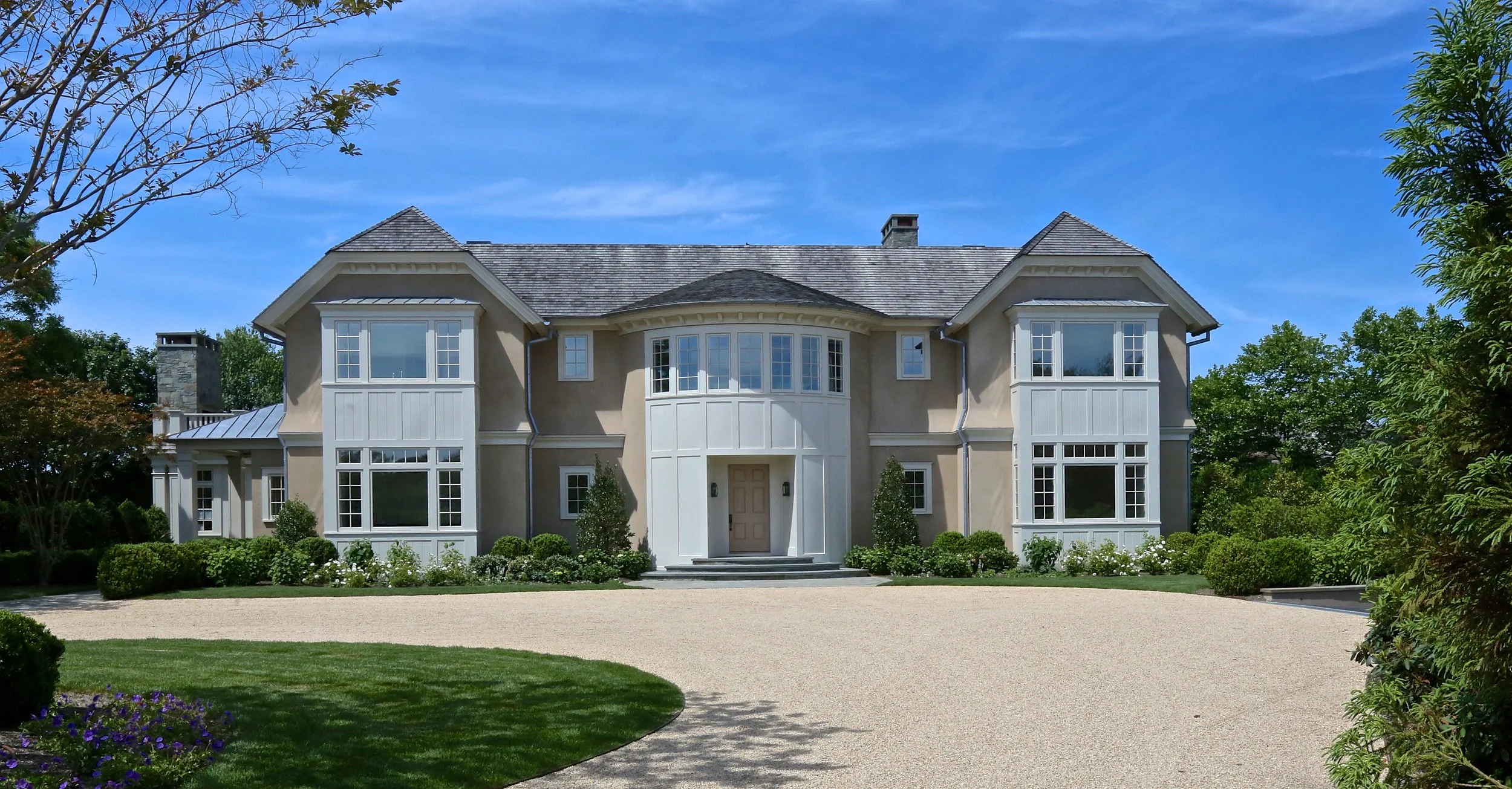 Large beige two-story house with white window frames and a central front door, surrounded by green bushes and trees, under a blue sky. Construction company in the Hamptons. 