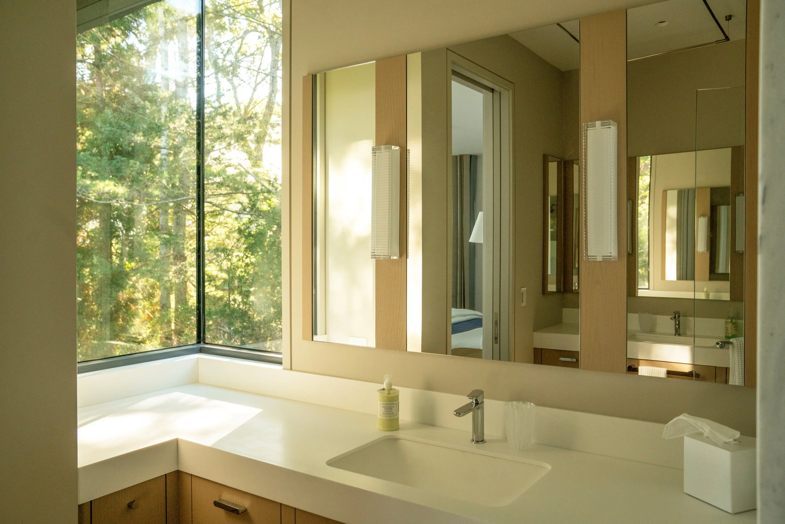 Modern bathroom with large window showing trees outside, white countertop with sink, hand soap, and tissue box, mirrors reflecting bedroom and other bathroom elements.