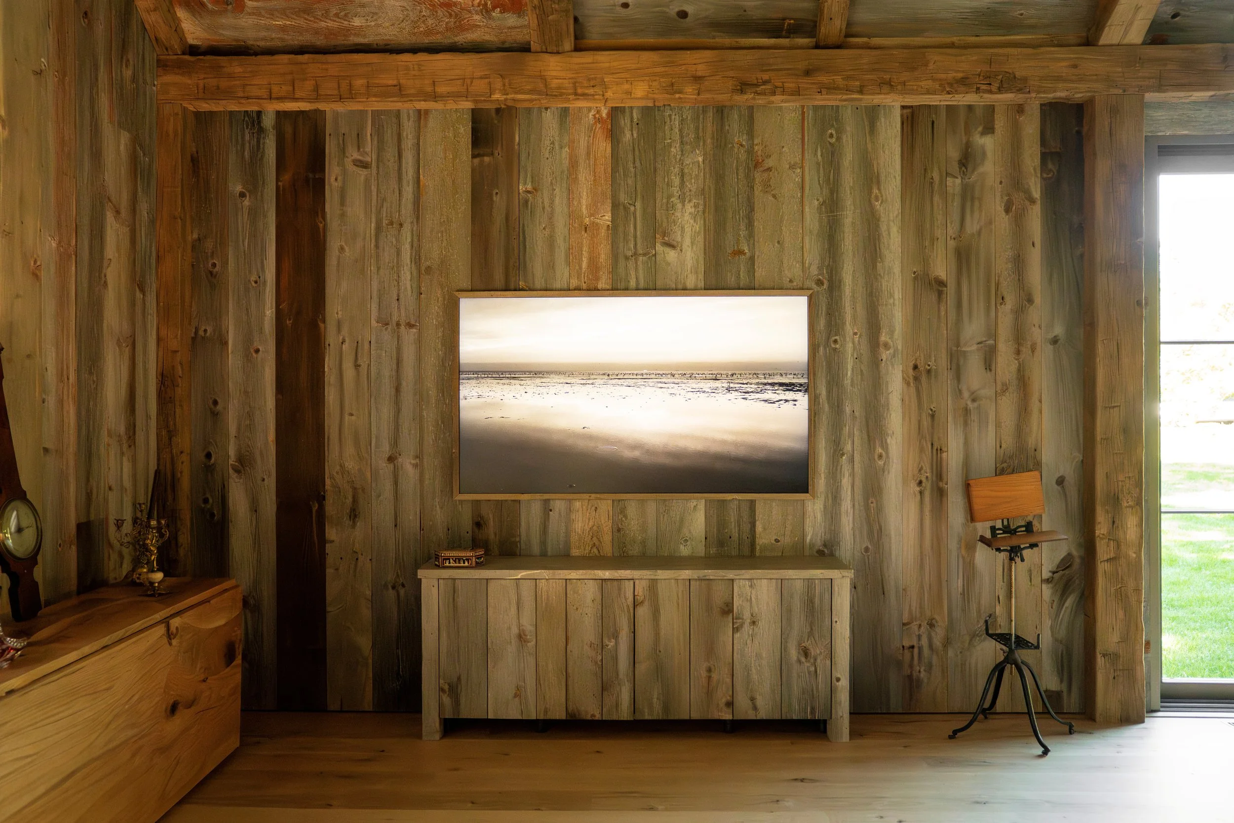 Interior of a rustic room with wooden wall paneling, a mounted landscape photograph of a beach at sunset, a wooden cabinet underneath, a small decorative item on top, and a vintage music stand near a window showing a green lawn outside.