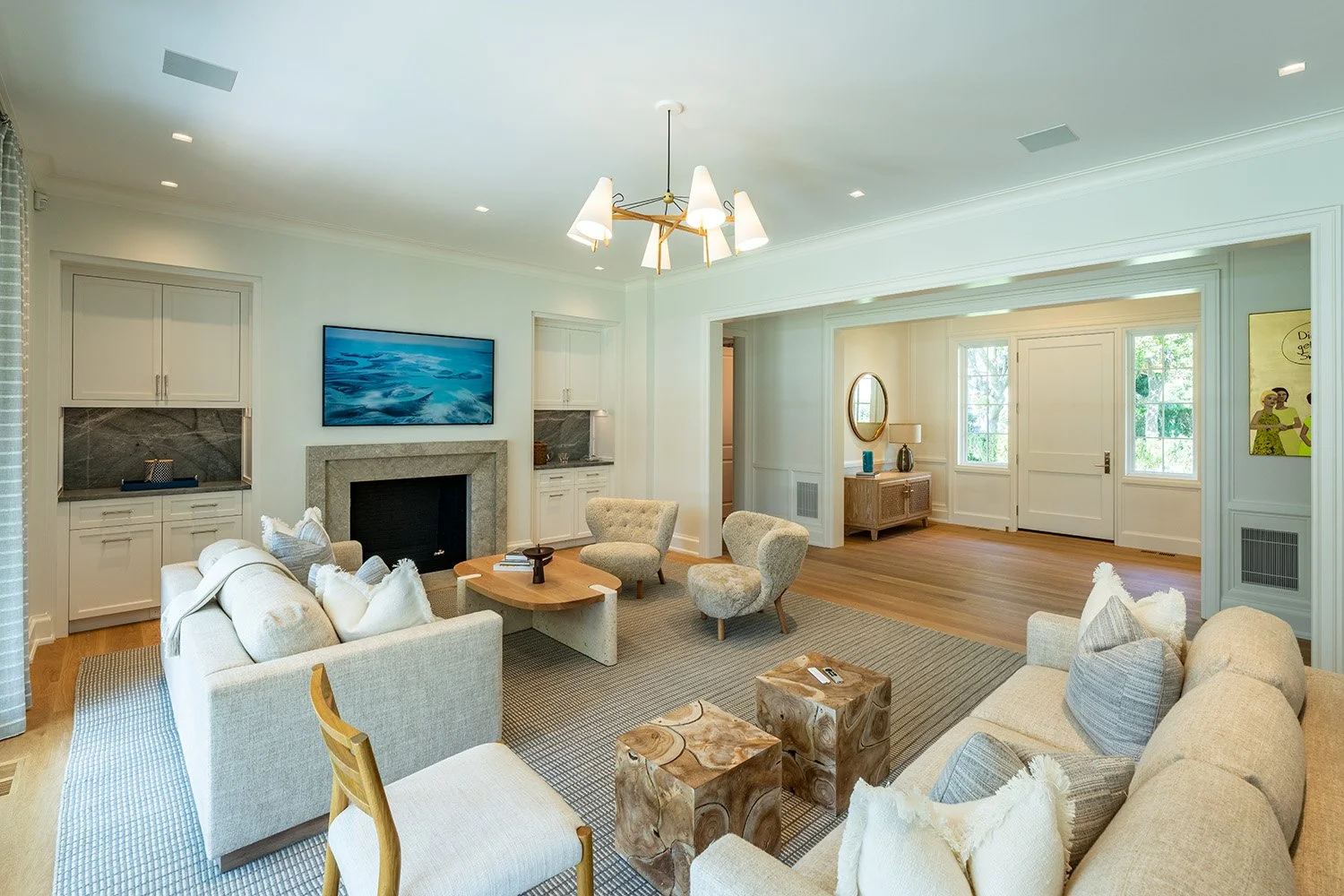 Living room with white walls, a stone fireplace with a TV above, beige sofas, and multiple chairs. Wooden side tables and a chandelier hang from the ceiling. Large windows let in natural light, and a wooden floor is partially covered by a rug.