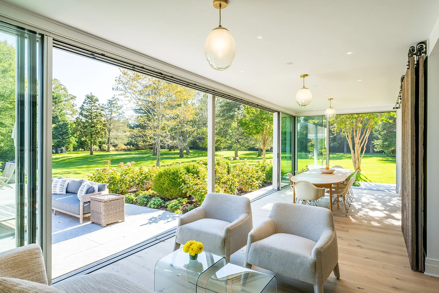 Bright living room with large glass sliding doors opening to a lush green garden, white armchairs, a glass coffee table with yellow flowers, and a dining table with chairs in the background.