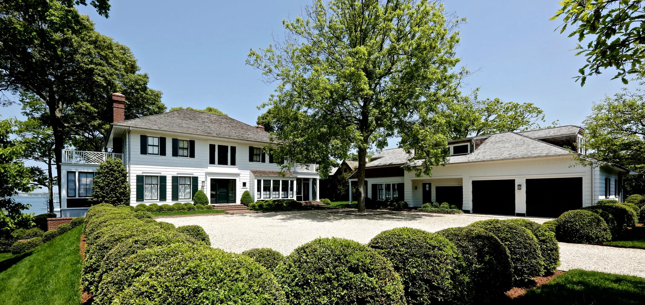 White two-story house with dark shutters, surrounded by trees, green shrubs, and a gravel driveway in a sunny setting.