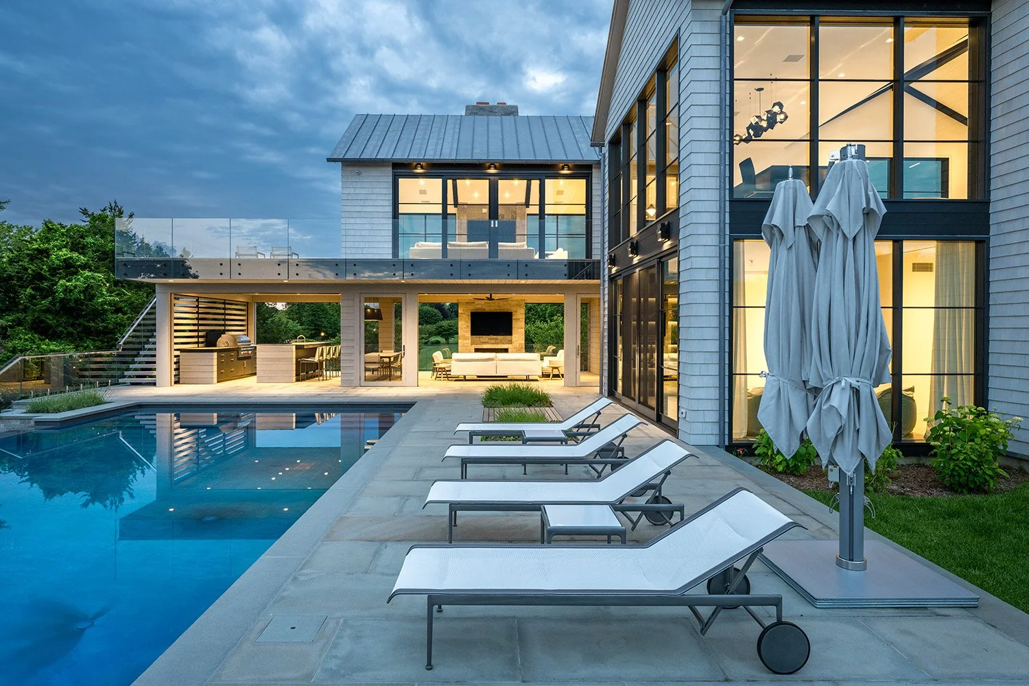 Modern house with large glass windows, outdoor pool, and poolside lounge chairs at dusk.