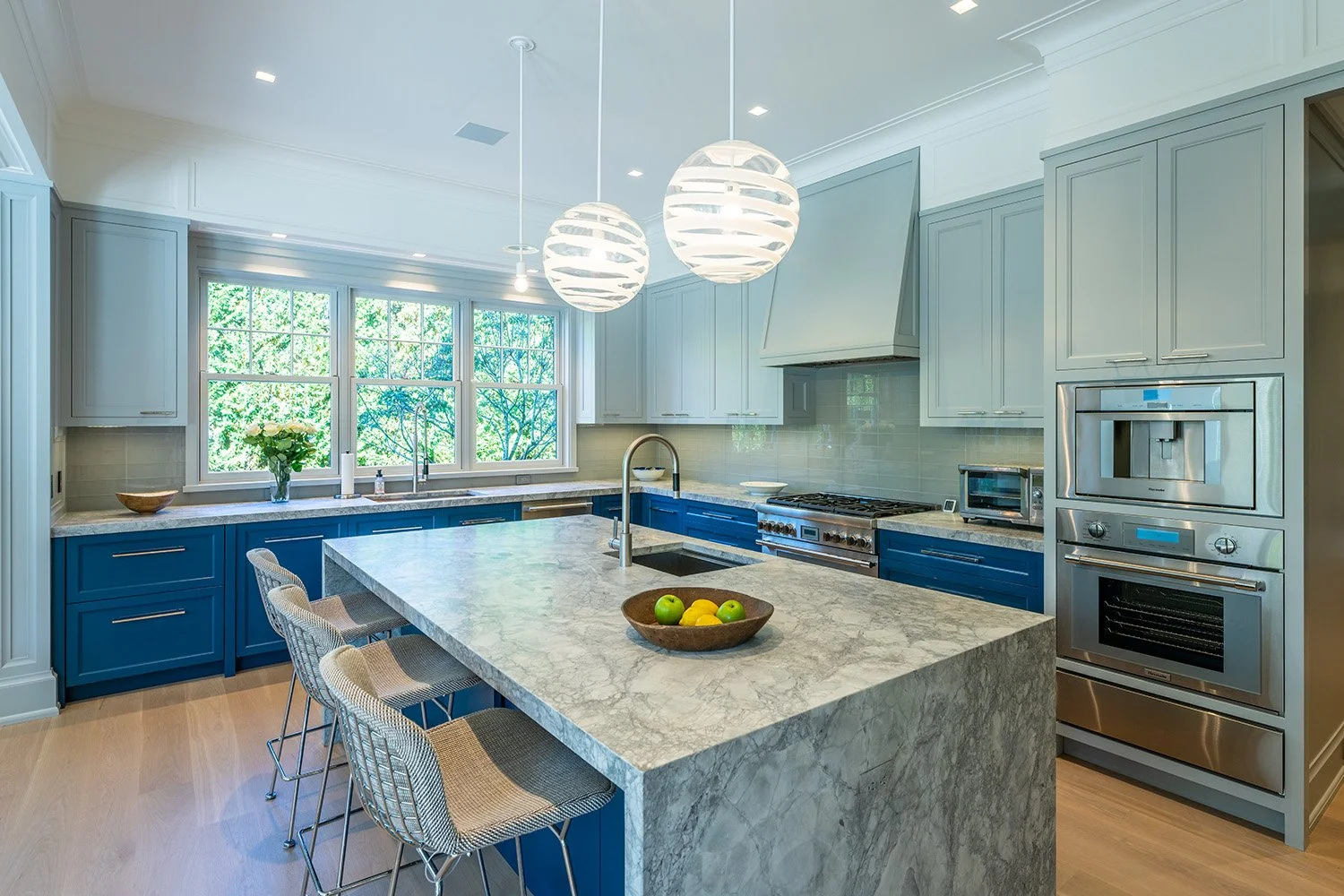 Modern kitchen with blue cabinets, white upper cabinets, marble island, large window, and stainless steel appliances.