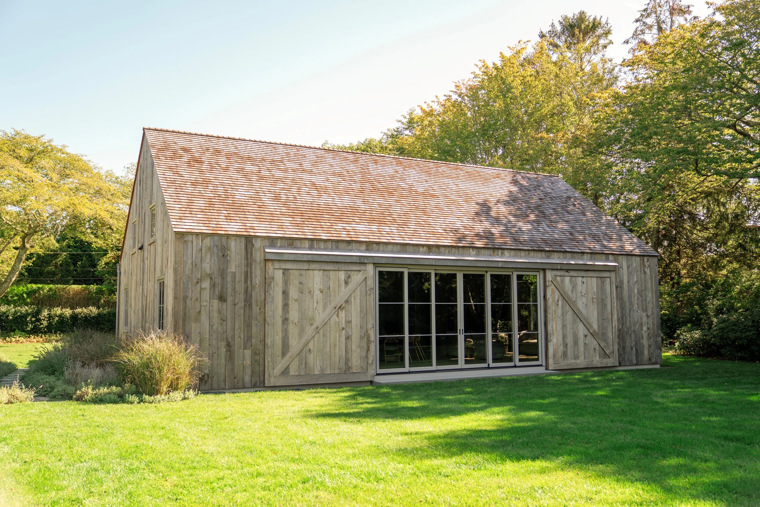 A wooden barn with a red shingled roof, set in a green grassy yard with trees in the background.