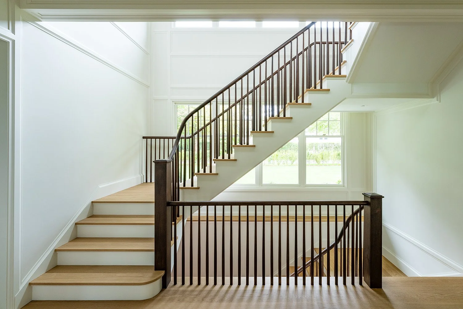 Interior view of a bright staircase with wooden steps, white walls, and large windows showing greenery outside.