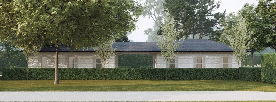 A single-story white brick building with a dark shingled roof, surrounded by green trees and a manicured hedge, with a lawn in the foreground. Hamptons Construction company.